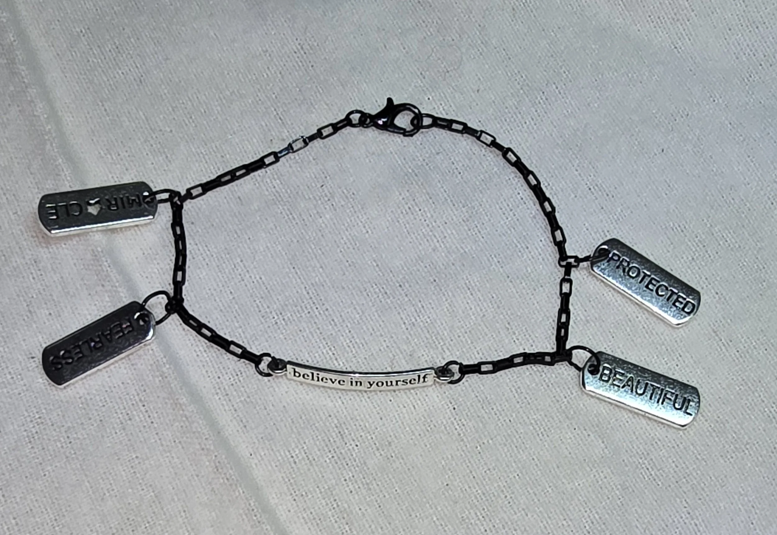 A black metal bracelet with five rectangular charms engraved with words: 'FEARLESS,' 'DREAM,' 'PROTECTED,' 'BEAUTIFUL,' and 'believe in yourself.'