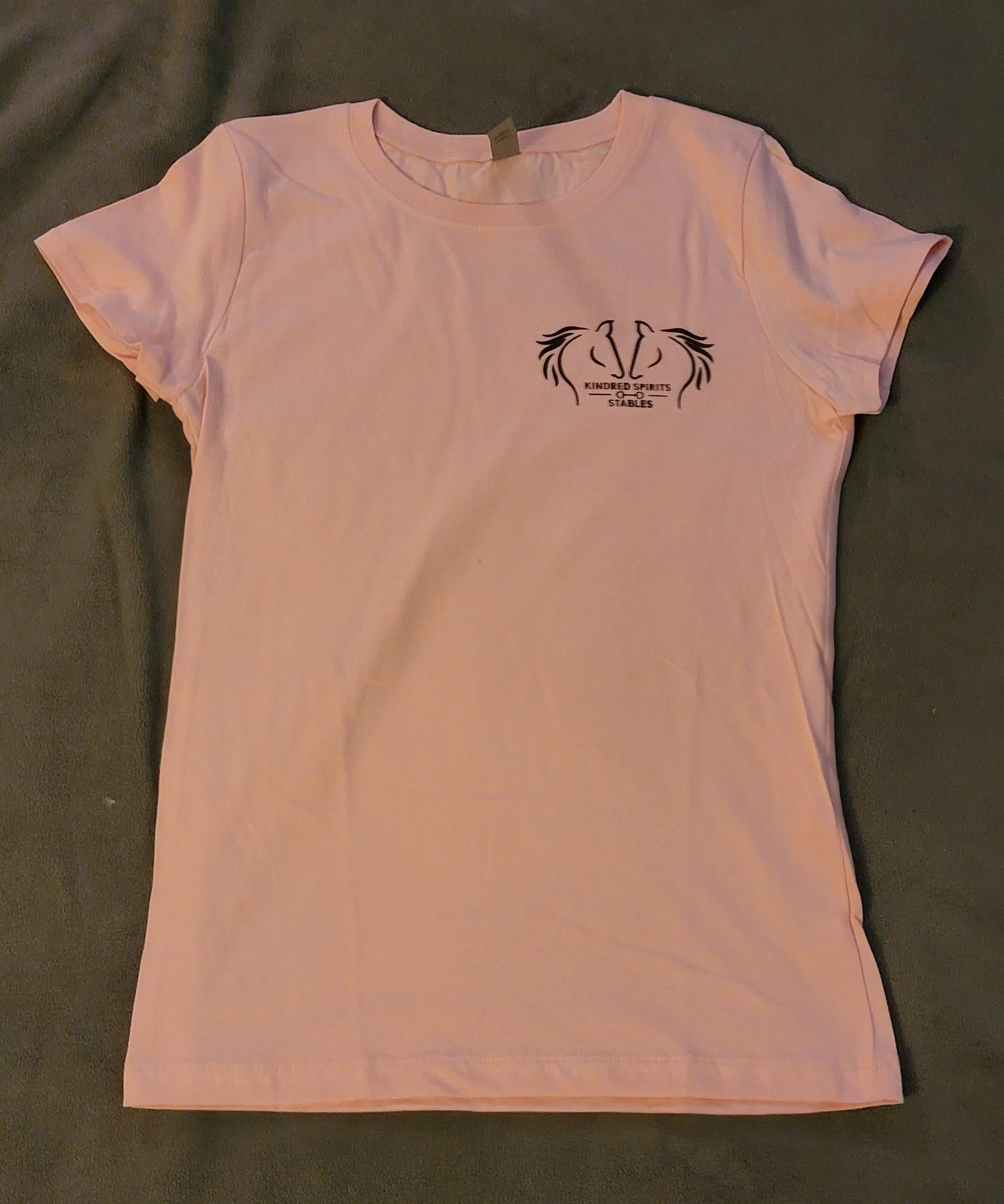 Light pink T-shirt with a small black logo on the left chest area, featuring two stylized horse heads facing each other with a line in between, and the words "Kindred Spirits Stables" underneath.