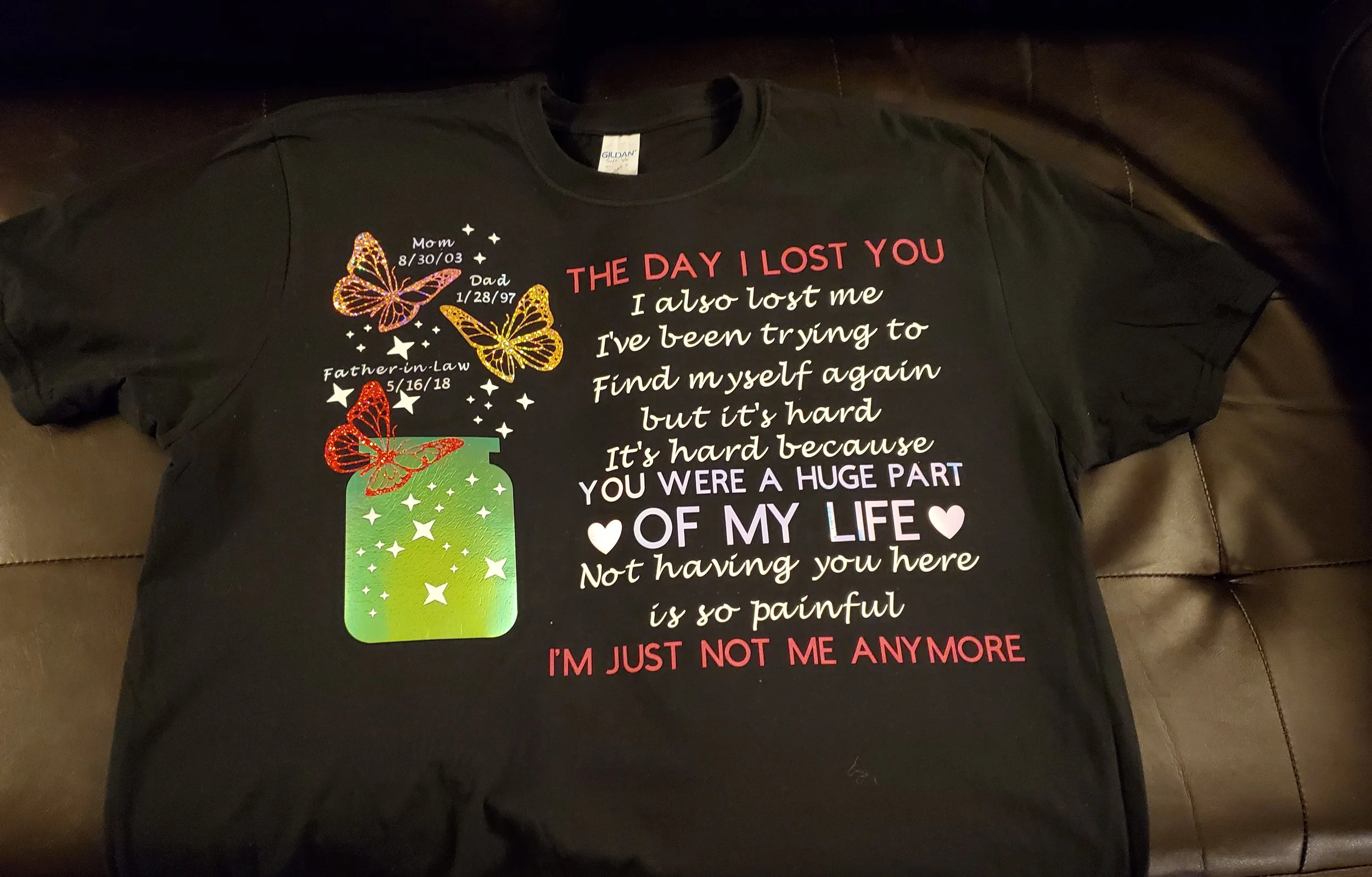 Black T-shirt with colorful text and graphics, including a green jar with white stars and three butterflies, one red, two yellow, and one purple, with dates and labels indicating family members and their birth dates.