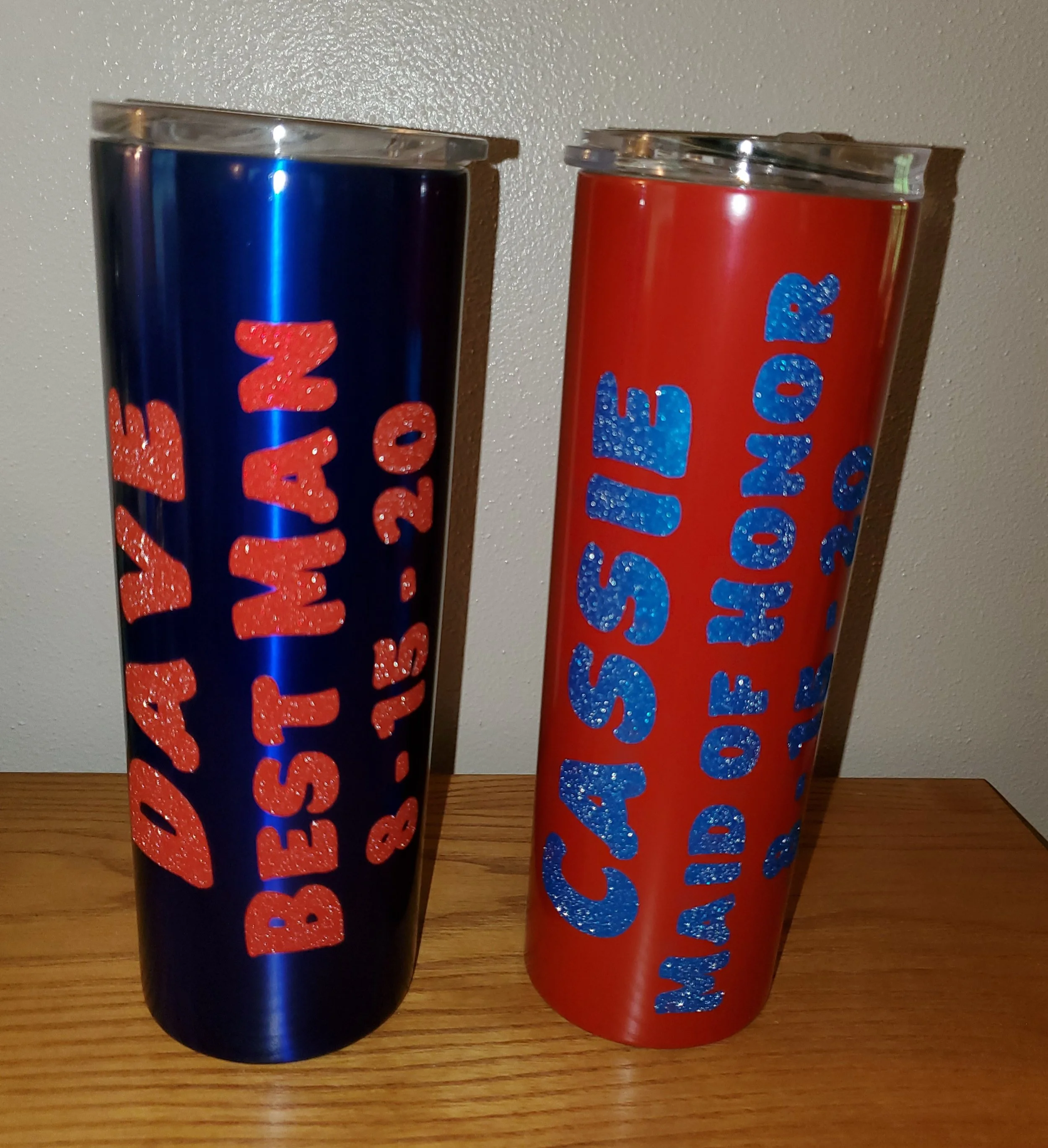 Two insulated tumblers with celebration themes, one in blue and one in red, placed on a wooden surface against a plain wall, with glittery text indicating Happy New Year and Cassie’s 21st birthday.