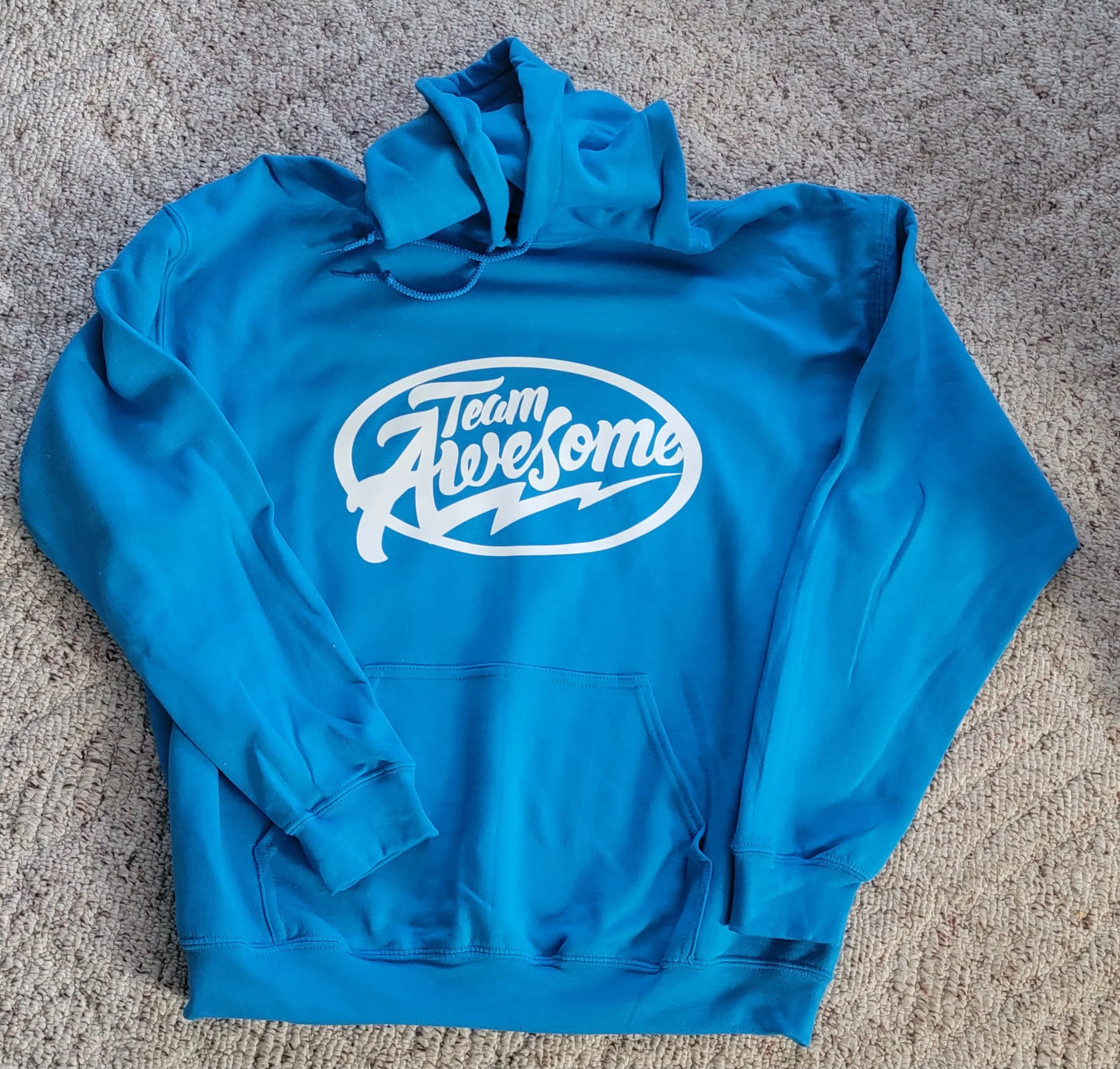 Blue hoodie with the text 'Team Awesome' in white with a swoosh design, laid on a beige carpet.
