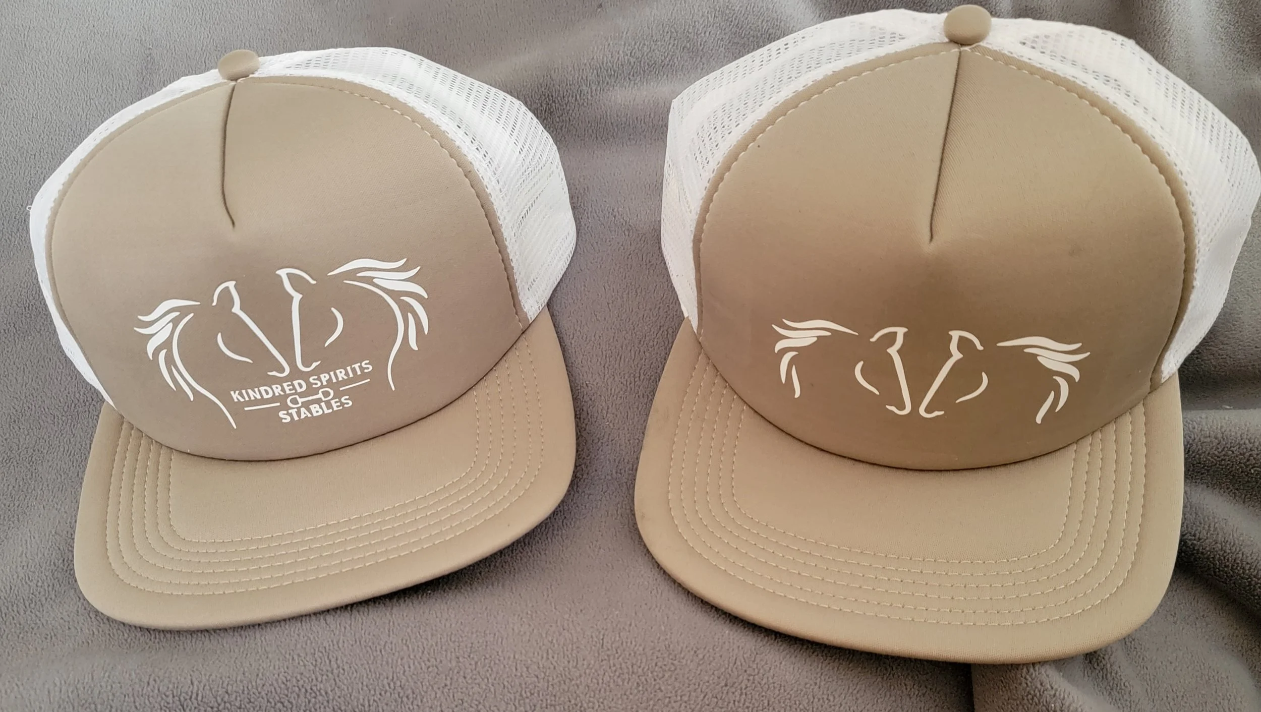 Two beige trucker hats with white mesh backing, featuring a logo of two stylized horses' heads facing each other, with the text 'Kindred Spirits Stables' underneath, placed on a gray surface.