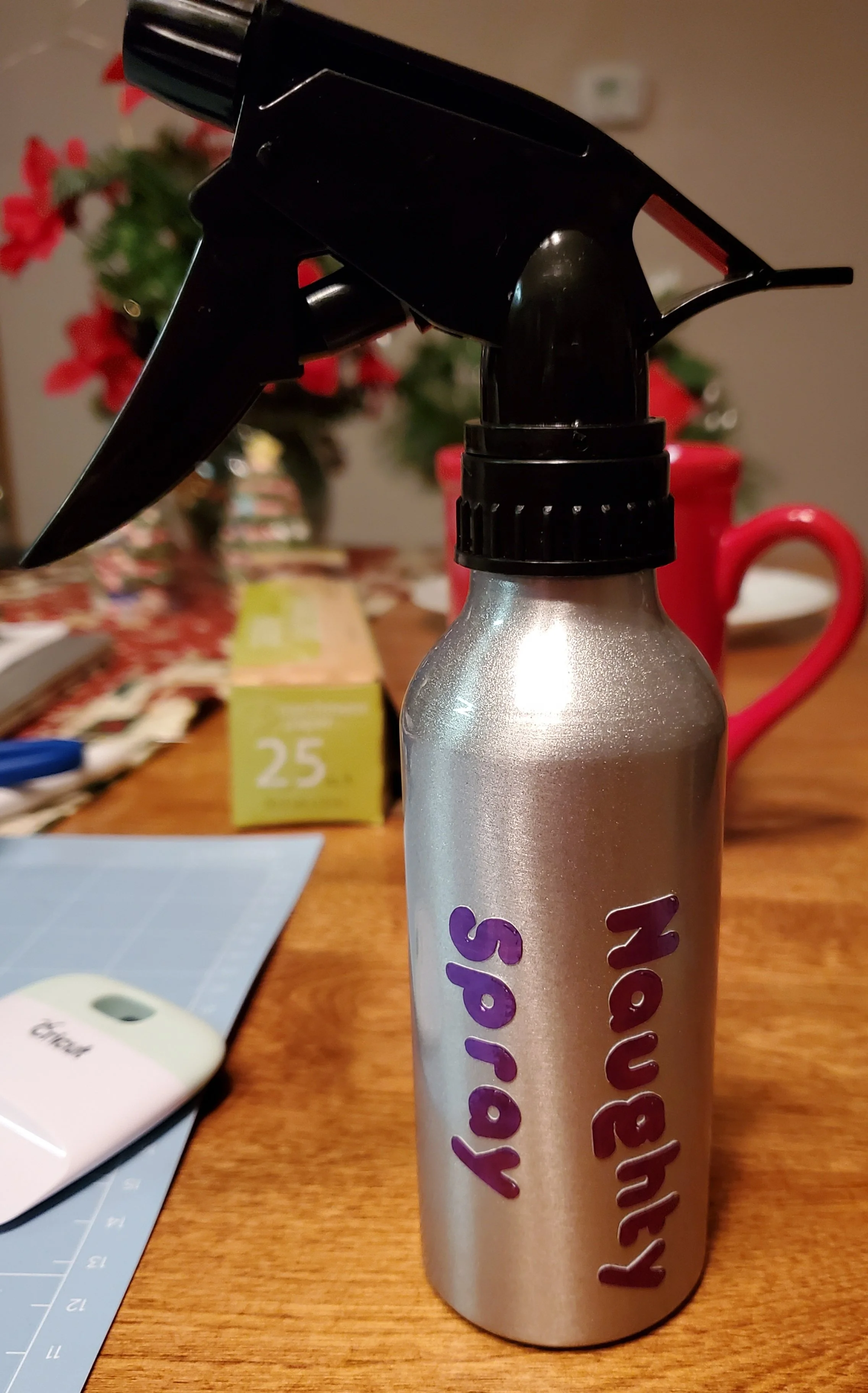 A silver spray bottle with a black nozzle on a wooden table, with a holiday-themed background including a poinsettia plant, red mug, and festive decorations.