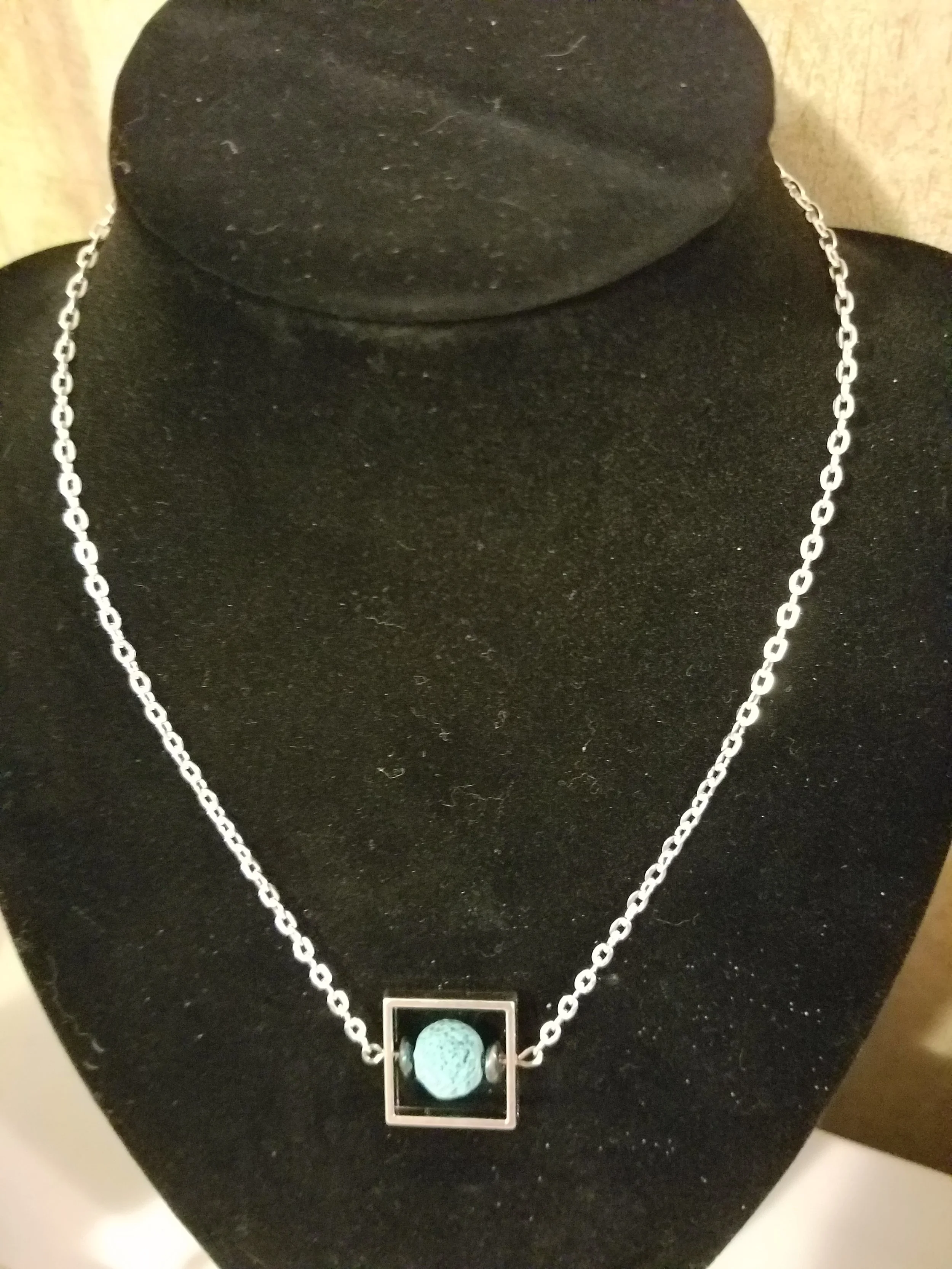 A silver chain necklace with a geometric square frame and a round turquoise pendant in the center, displayed on a black velvet bust.