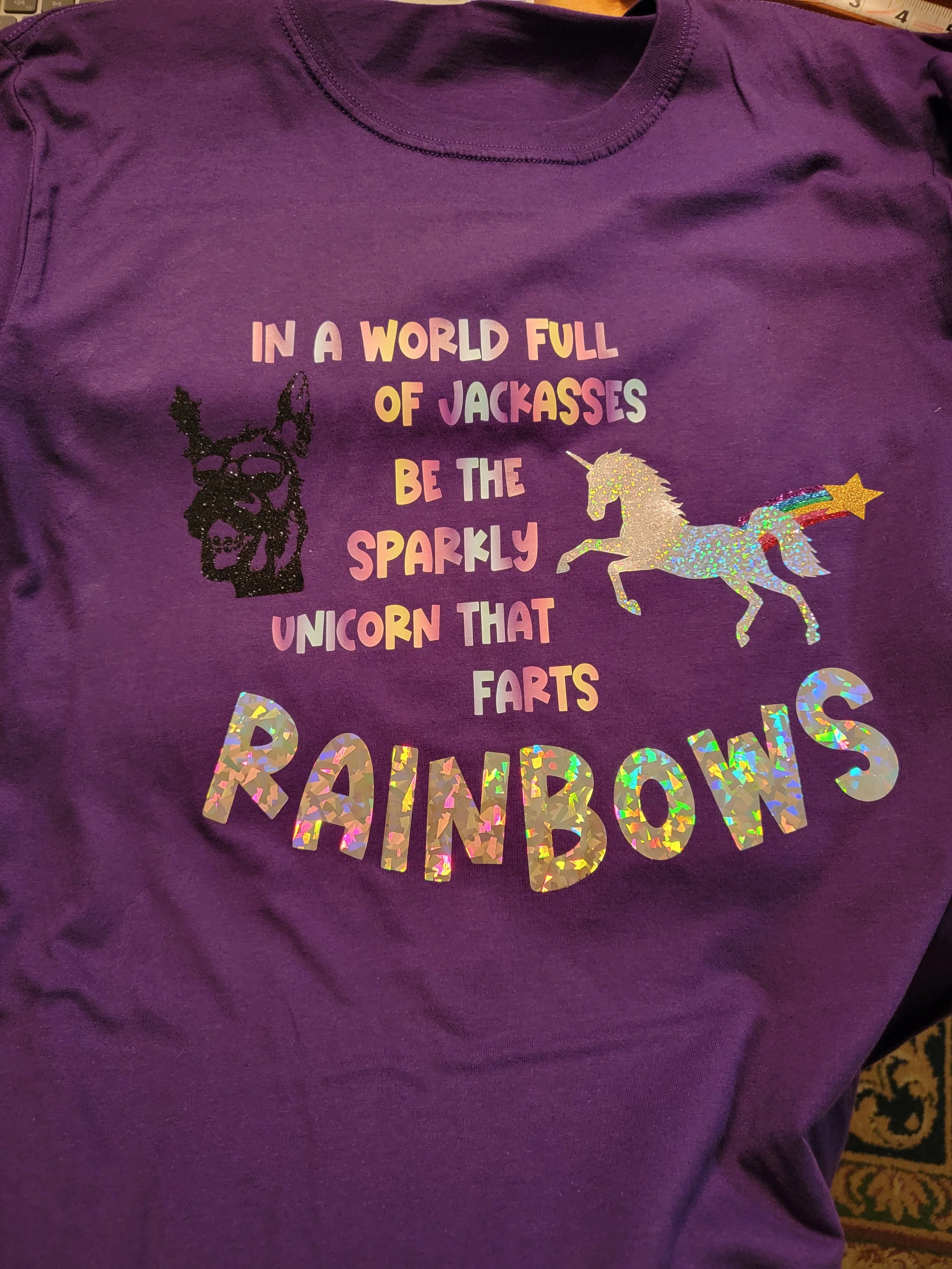 Purple T-shirt with colorful text and illustrations. The text reads: 'In a world full of jackasses be the sparkly unicorn that FARTS rainbows.' The shirt features a black glittery dog face, a glittery unicorn, and glittery rainbow-colored text spelli