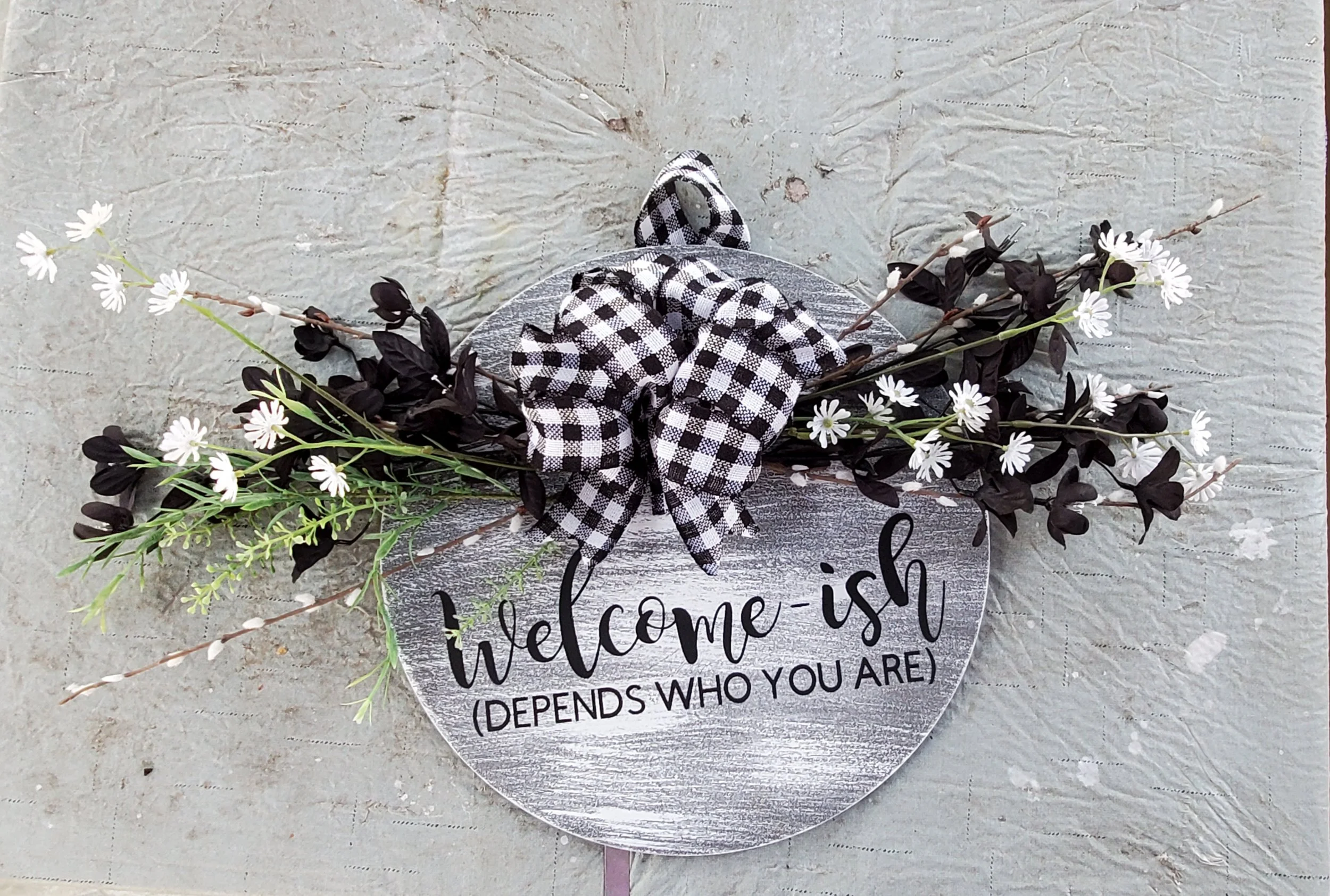 A decorative welcome sign with white and black gingham bow and artificial white and purple flowers, on a textured light gray surface.