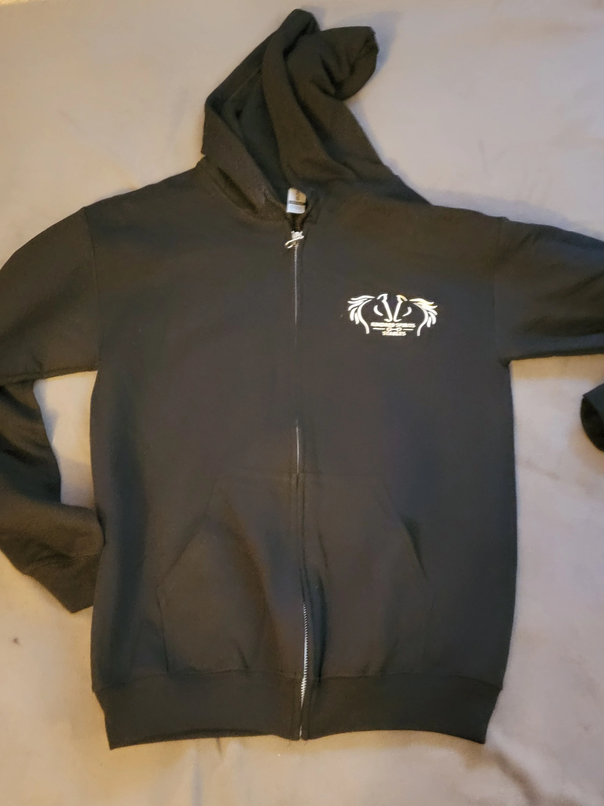Black zip-up hoodie with a white crest and decorative design on the left chest, lying on a beige surface.