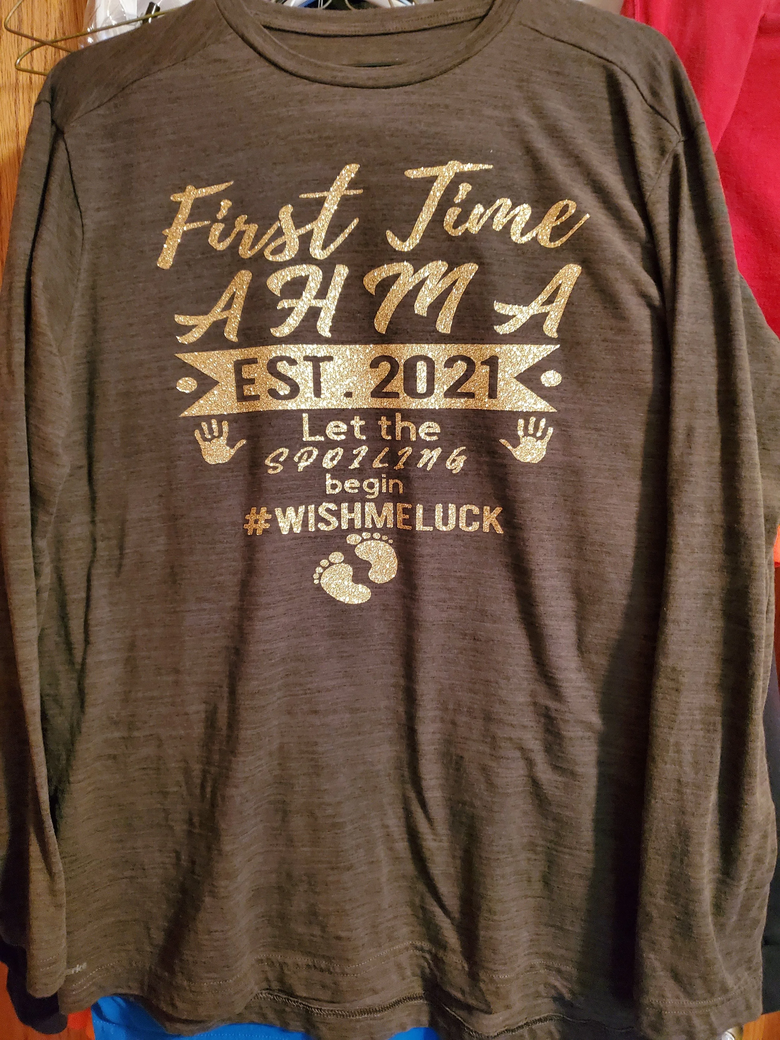 Long sleeve shirt with gold-colored glitter text that reads, 'First Time A H M A EST. 2021 Let the Spilling begin #WISHMELUCK' and features a pair of baby footprints.