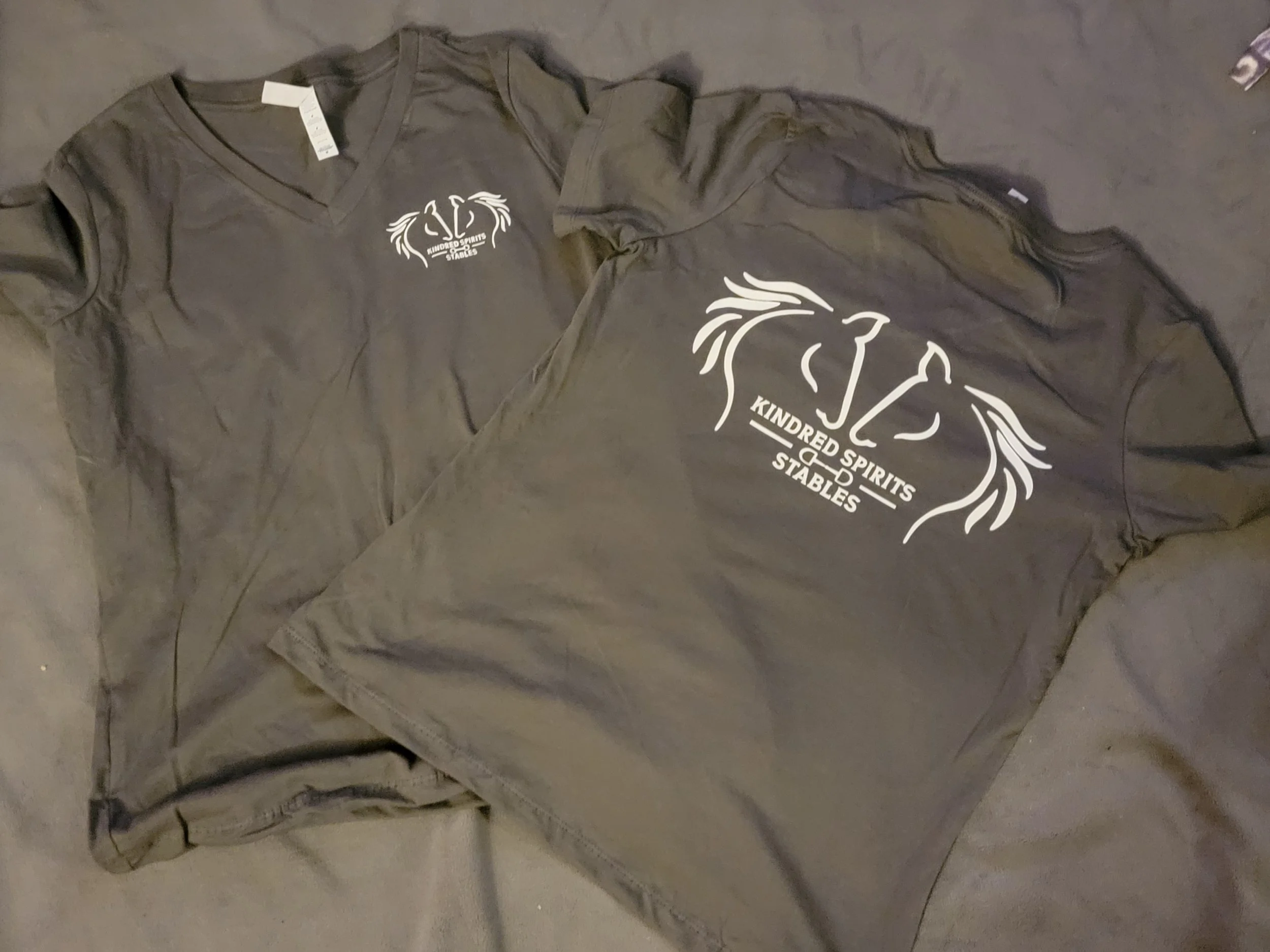 Gray t-shirt and gray hoodie with a white logo and text that reads 'Kindred Spirits Stables' on a gray surface.