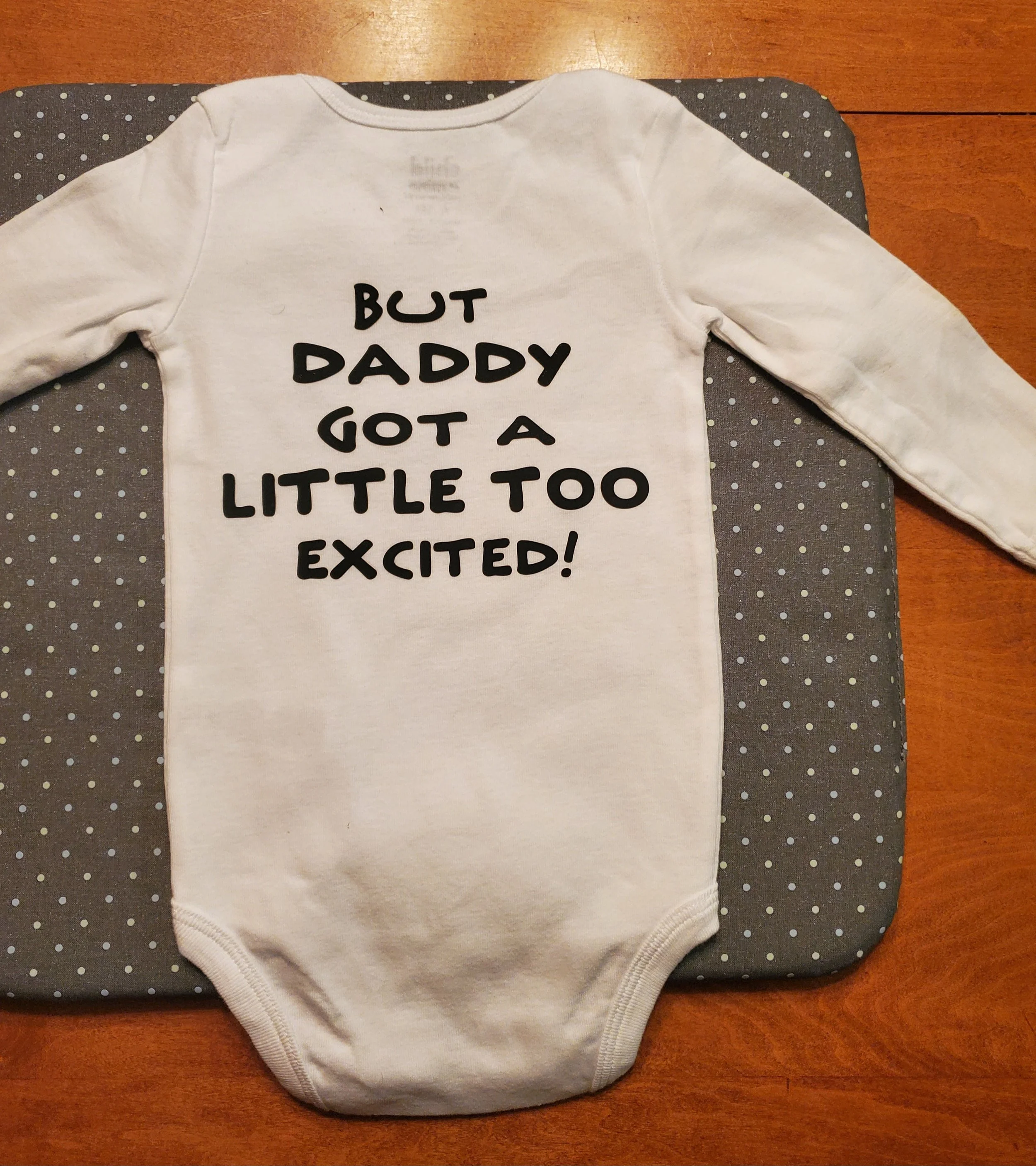 White baby onesie with black text that says, 'But Daddy Got a Little Too Excited!', laid flat on a changing pad with a polka dot pattern on a wooden surface.