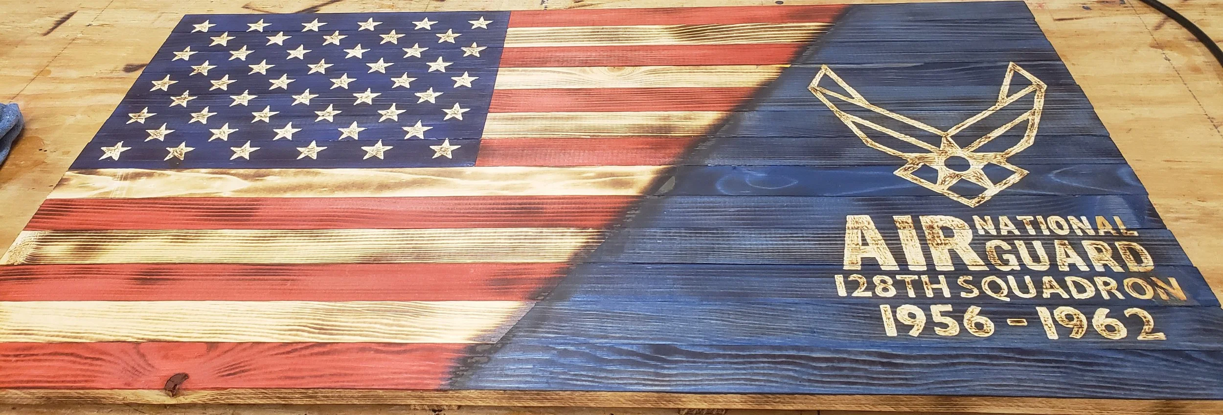 A wooden American flag with a blue field of white stars on the left and red and white stripes on the right, painted on wood. The right side features a gold Air Guard emblem and text dedicated to the 128th Squadron, 1956-1962.