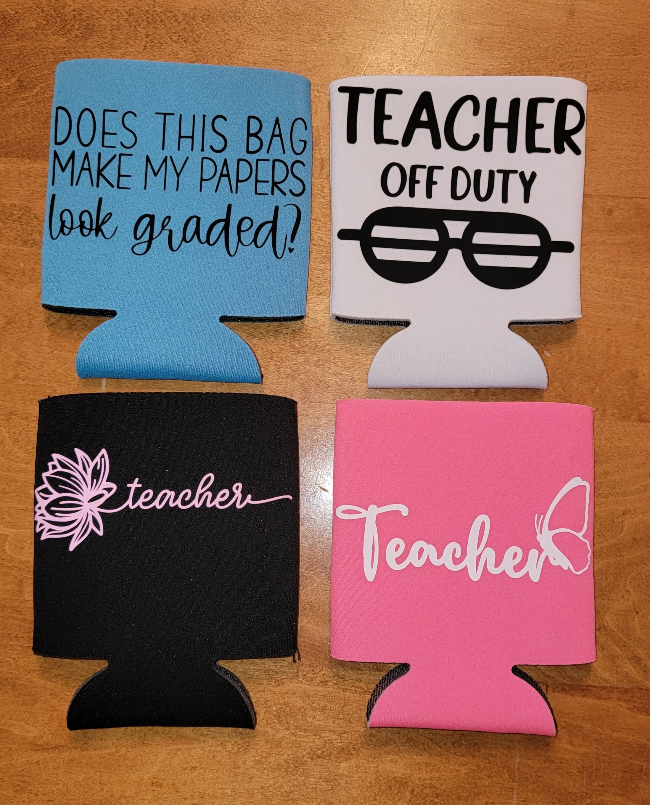 Four foam drink toppers with sayings and graphics related to teachers, placed on a wooden surface. The top left blue topper reads, 'Does this bag make my papers look graded?' The top right white topper says, 'Teacher off duty' with a graphic of glass