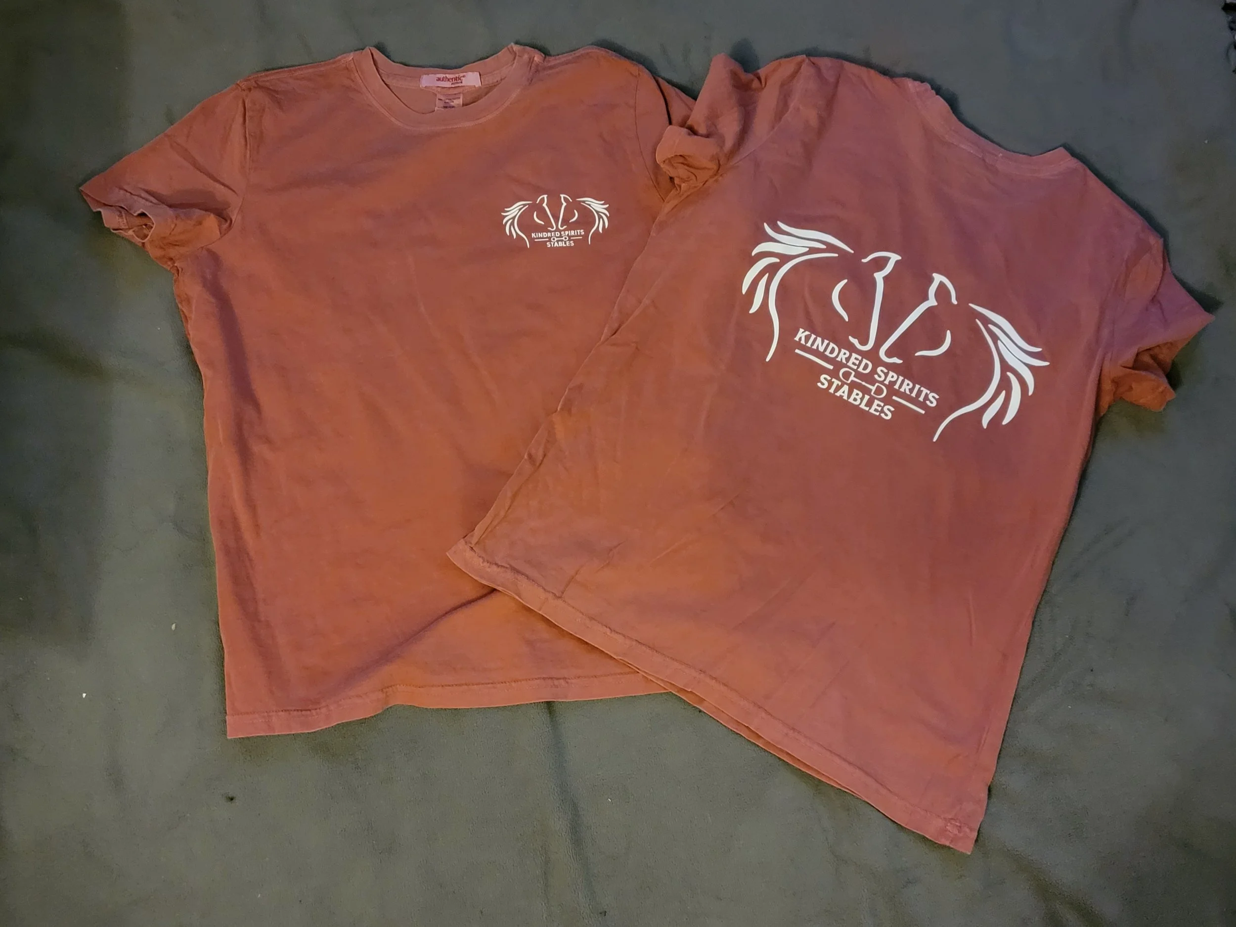 Two maroon T-shirts laid flat on a dark surface. The T-shirts feature a white logo with stylized horse heads and the text "KINDRED SPIRITS STABLES" underneath.