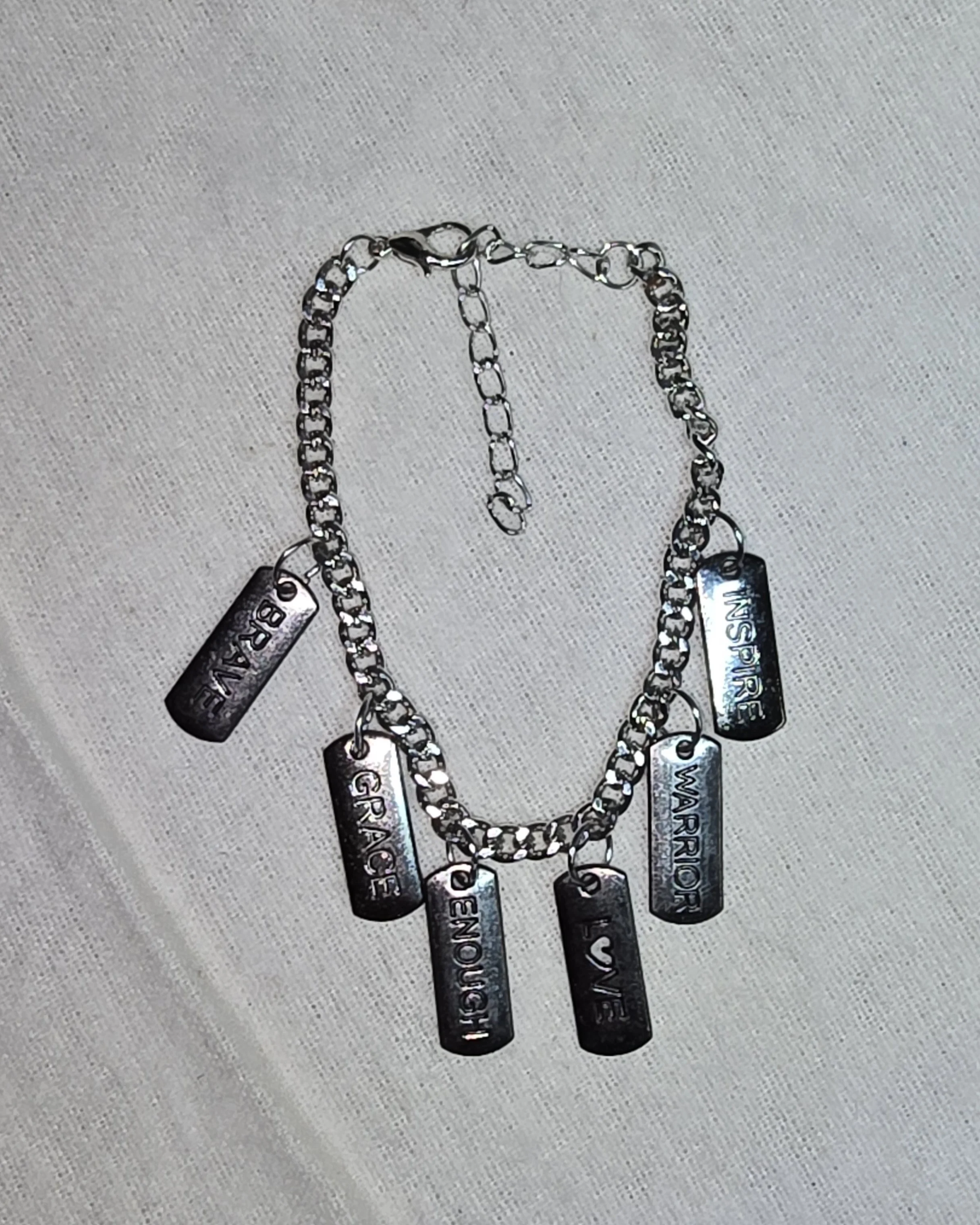 A silver chain necklace with six rectangular charms engraved with words 'GRACE', 'LOVE', 'VIA', 'WARRIOR', 'INSPIRATION', 'BELIEVE' and a small heart symbol.