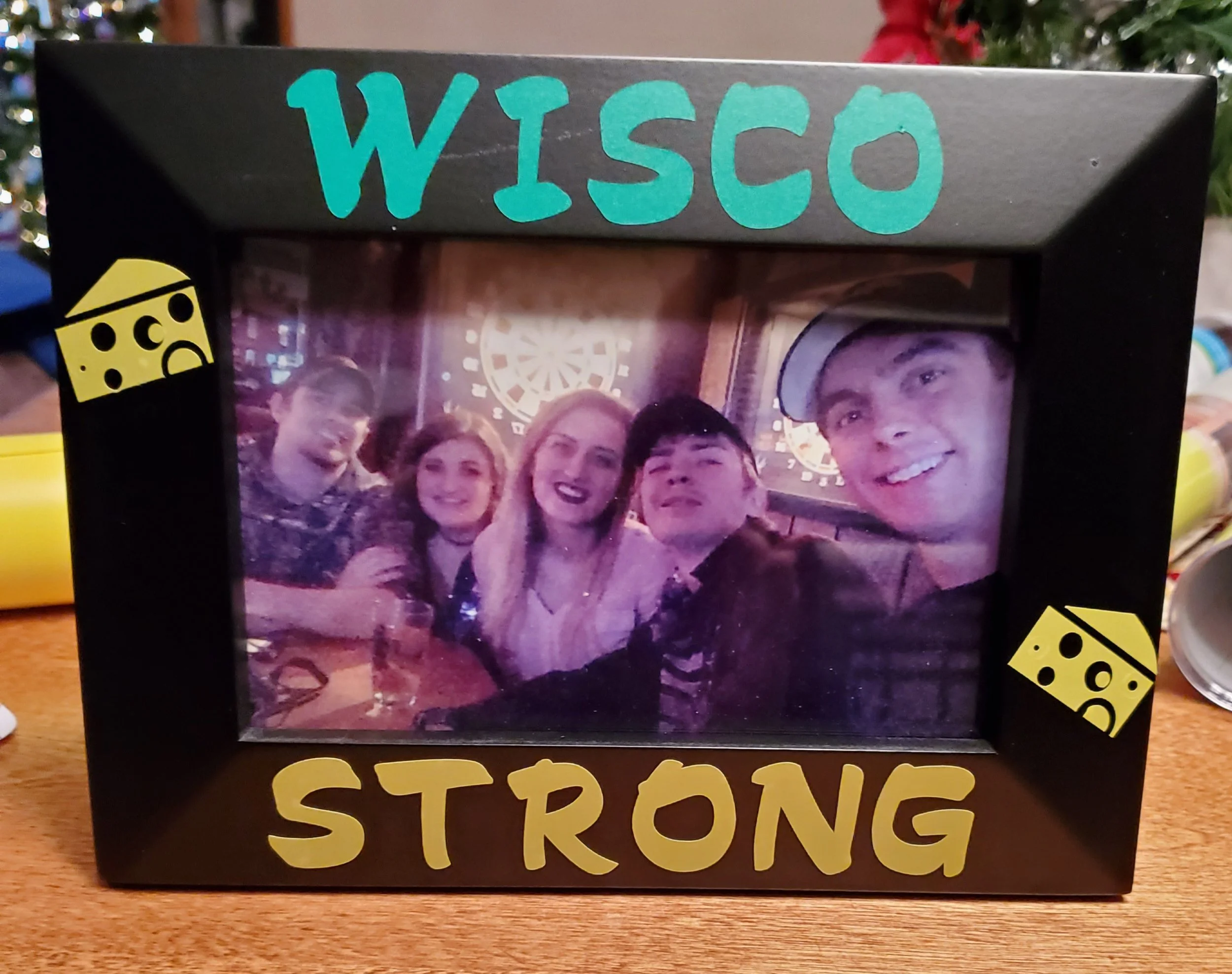 Photograph of five young adults smiling at the camera inside a black picture frame. The frame is decorated with yellow and teal dice and the words 'WISCO' at the top and 'STRONG' at the bottom. The background shows a bar or arcade setting with a dart