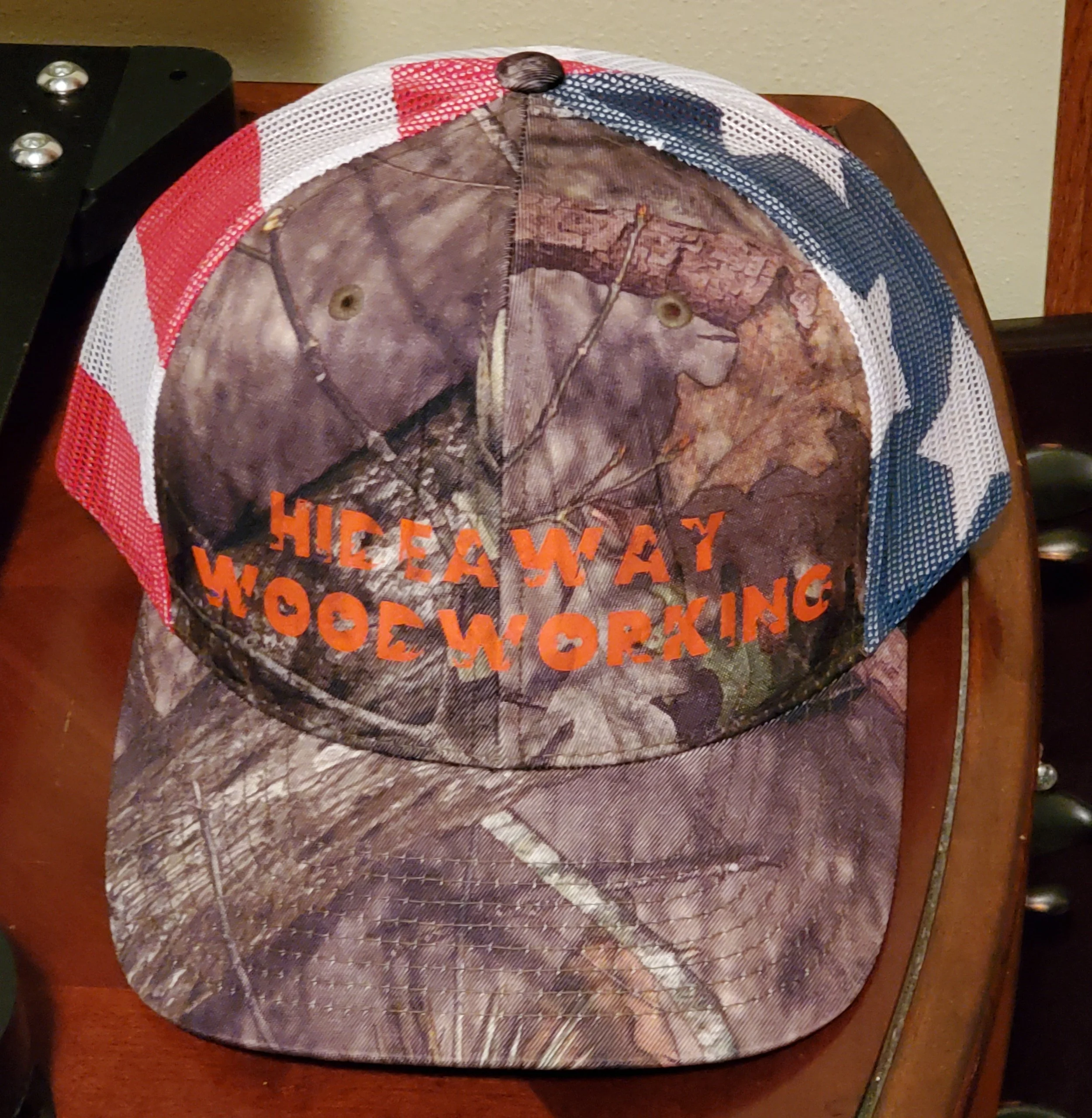 Camouflage baseball cap with orange text that reads 'HIDEAWAY WOODWORKING' and patriotic red, white, and blue mesh panels on the sides.