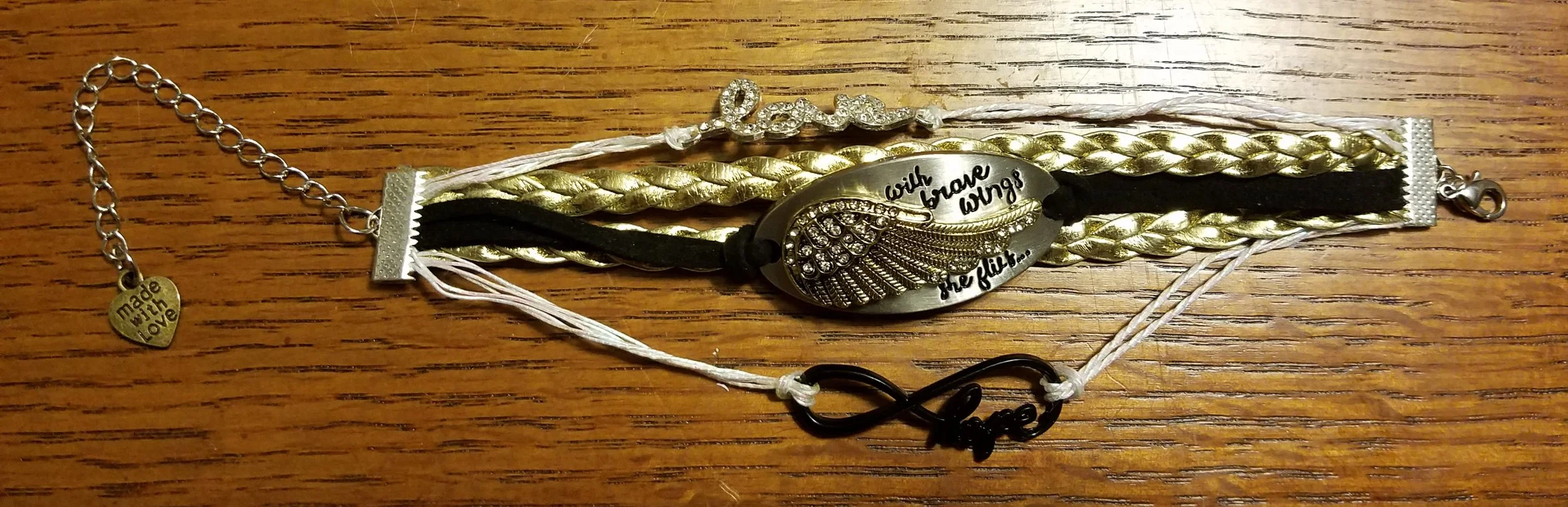 Bracelet with multiple cords, a metal oval pendant with a wing design and words, a heart-shaped charm with 'made with love' engraved, and an infinity symbol with the word 'love' on a wooden surface.