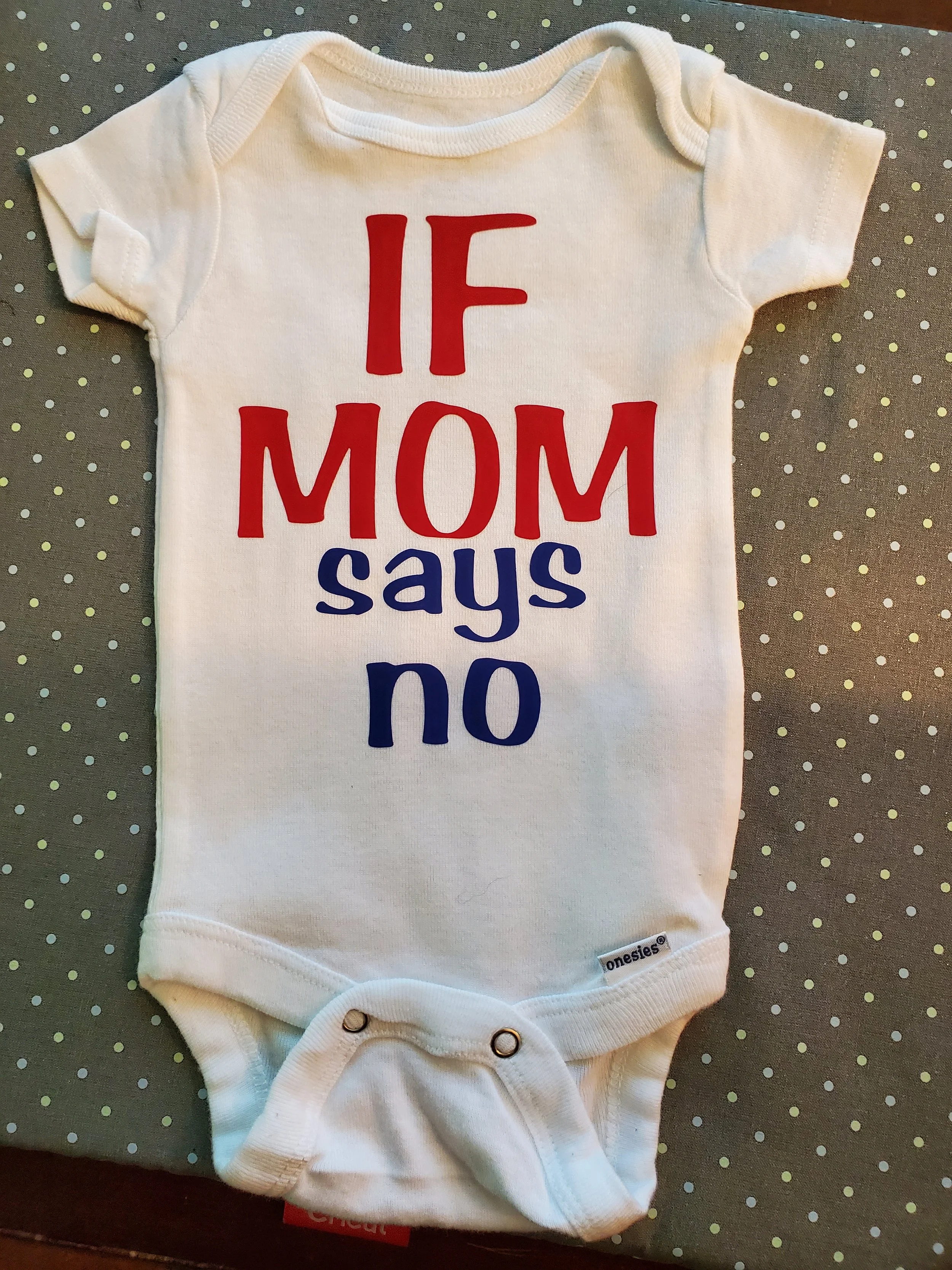 A white baby onesie with the printed phrase 'IF MOM says no' in red and blue letters. The onesie is laid flat on a gray polka dot surface.