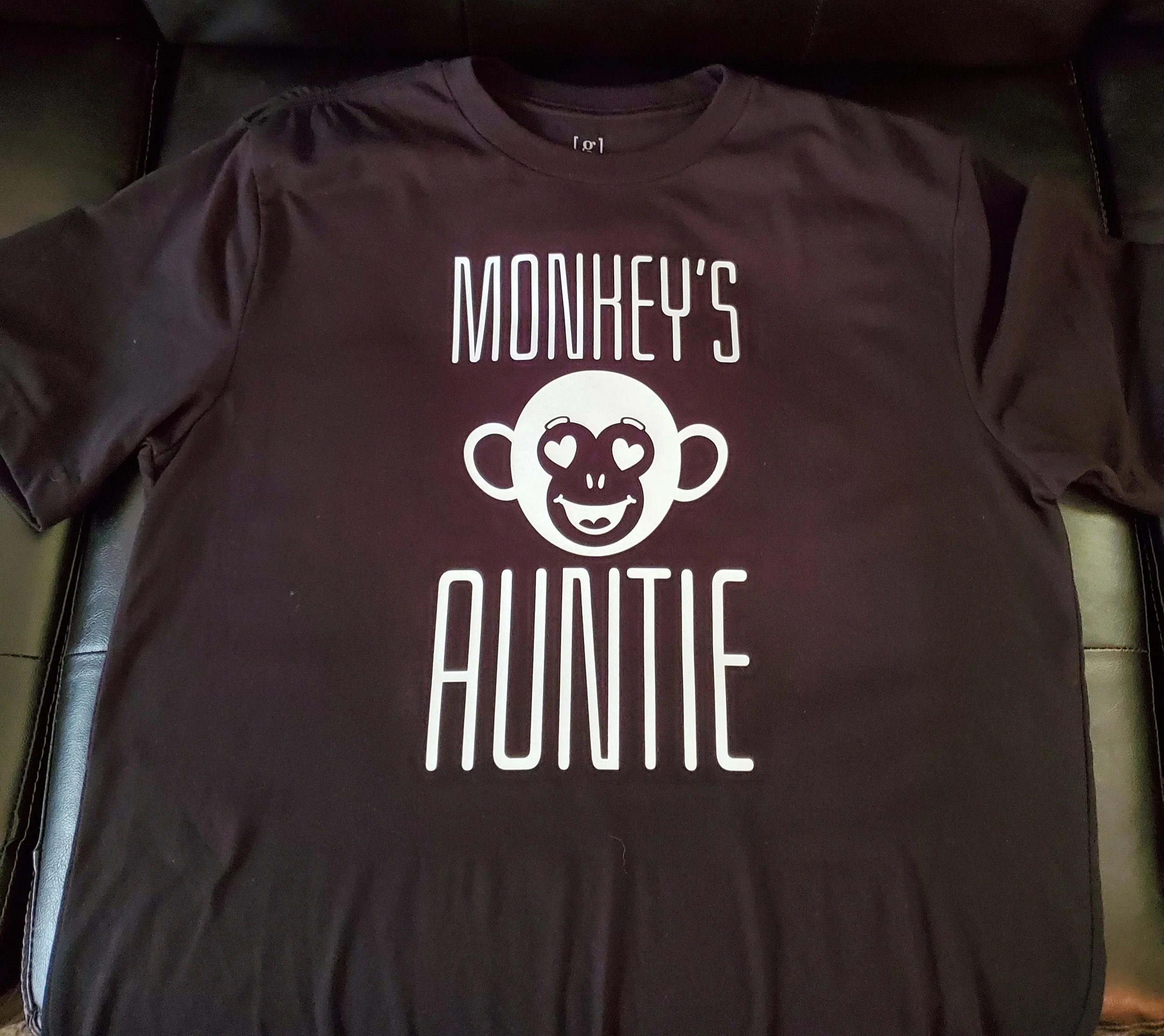 Black T-shirt with white text and a monkey face graphic. The text says 'Monkey's Auntie' with the monkey face showing heart-shaped eyes and a smiling mouth.