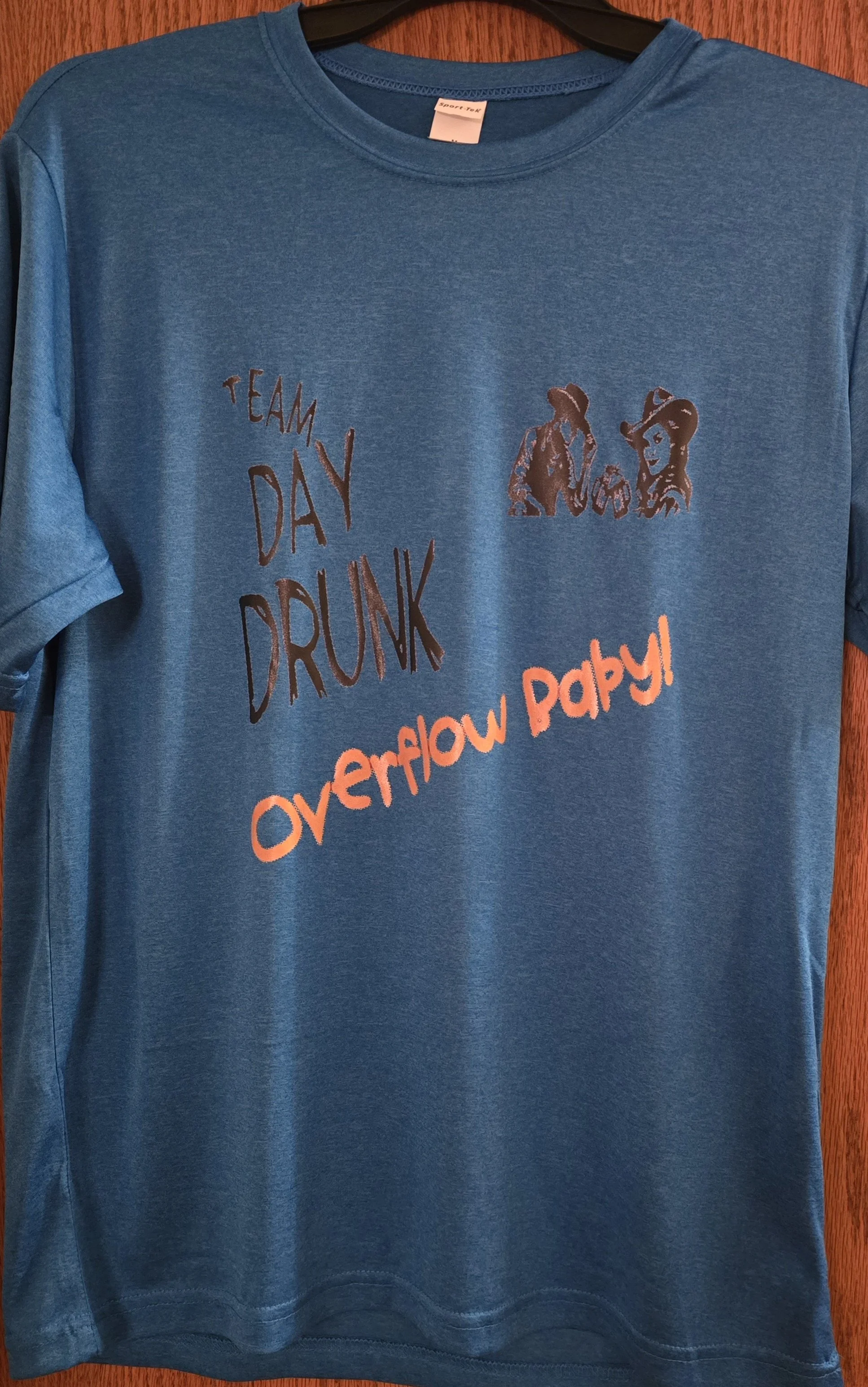 Blue T-shirt hanging on a black hanger with printed text and graphics, featuring the phrase "Team Day Drunk" and "Overflow Party" in orange, and an illustration of two women in cowboy hats.