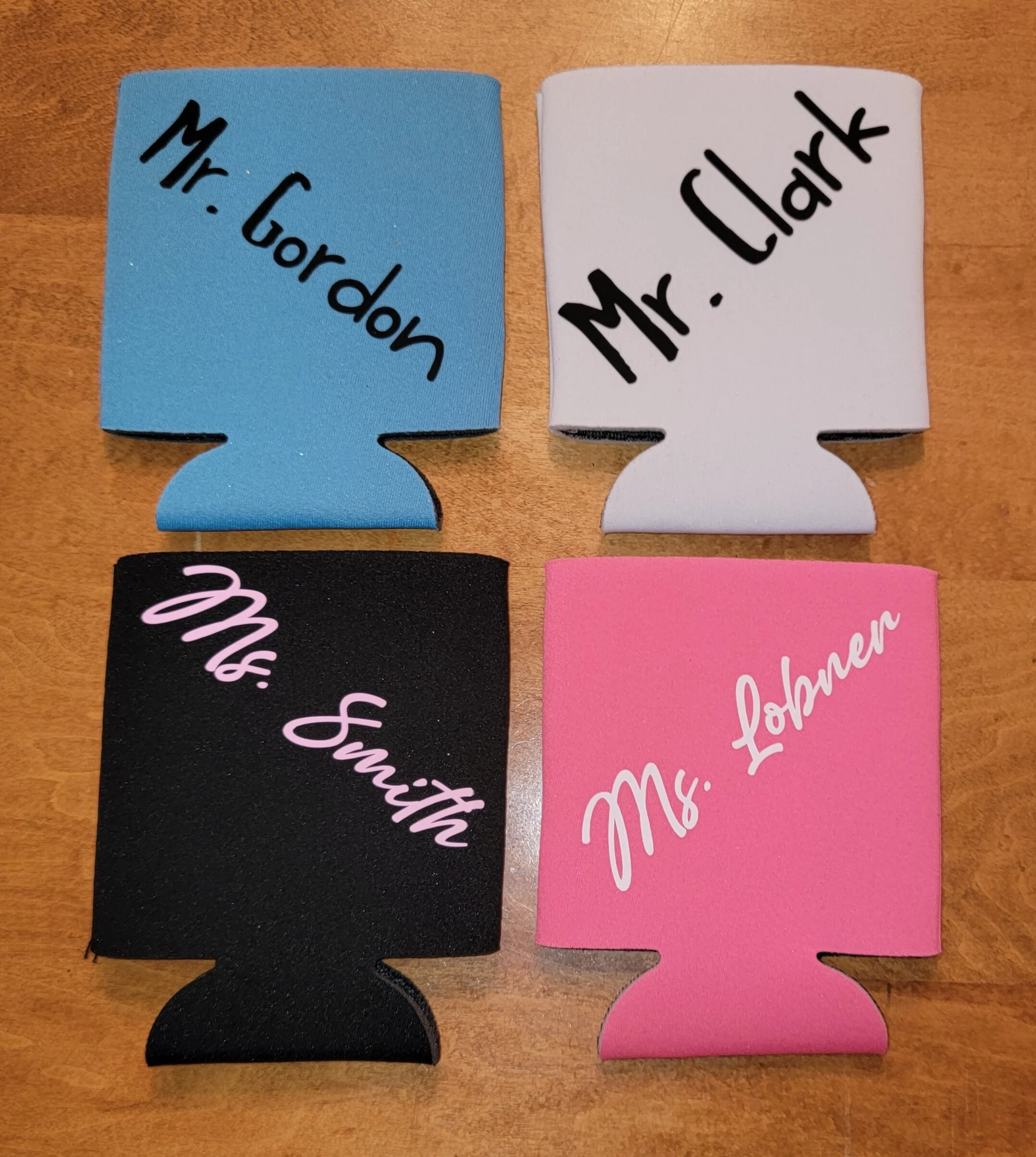 Four can cooler koozies in blue, white, black, and pink with printed names 'Mr. Gordon', 'Mr. Clark', 'Mr. Smith', and 'Mr. Coben' on a wooden surface.