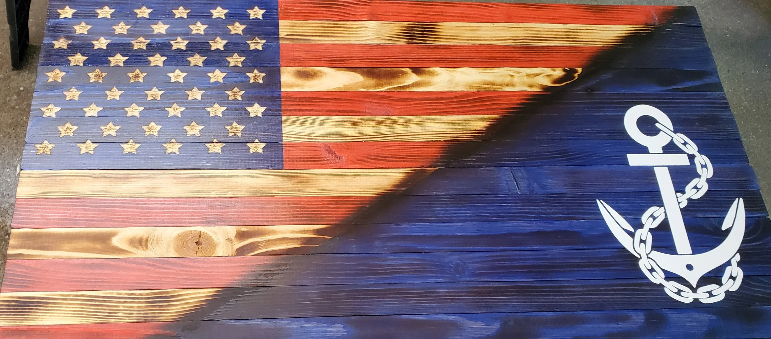 Wooden surface painted with the American flag. The flag features red and white stripes and white stars on a blue field. A white anchor with a chain is painted on the bottom right corner.