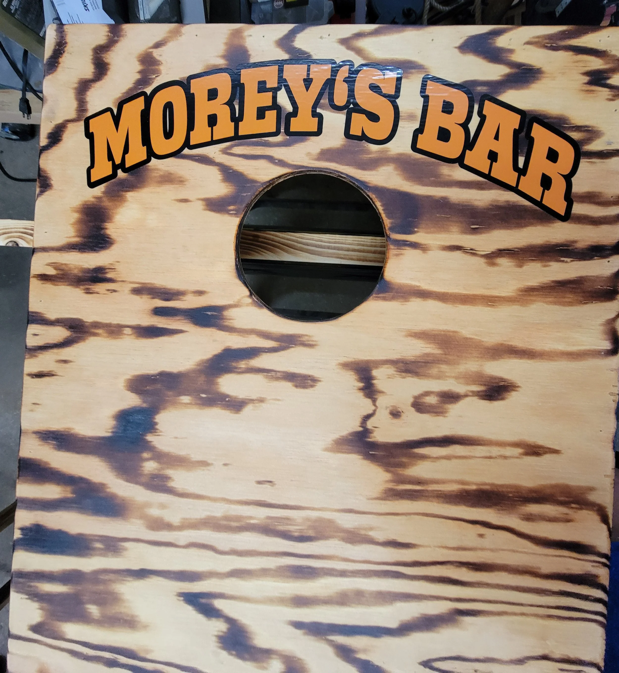 A wooden cornhole game board with a circular hole in the center and the words 'Morey's Bar' written on it in bold orange and black. The board has a burnt wood pattern design.