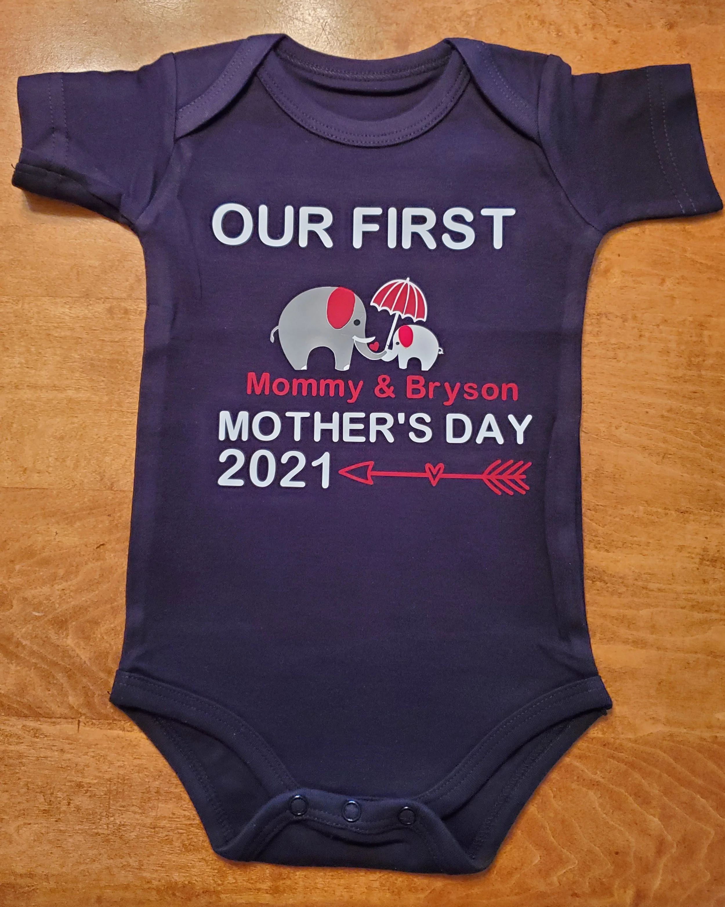 Baby Onesie in dark blue with a design for Mother's Day 2021. Features two elephants, one large and one small, holding an umbrella. The elephants are gray with red ears, and the umbrella is red and white. Text on the onesie reads: 'Our First Mommy & 