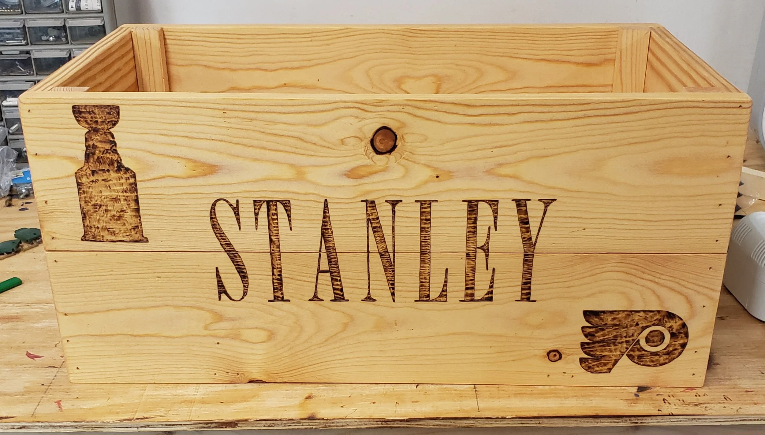 A wooden Stanley toolbox with the Stanley logo and two stamped logos, one on each side, resting on a workbench.