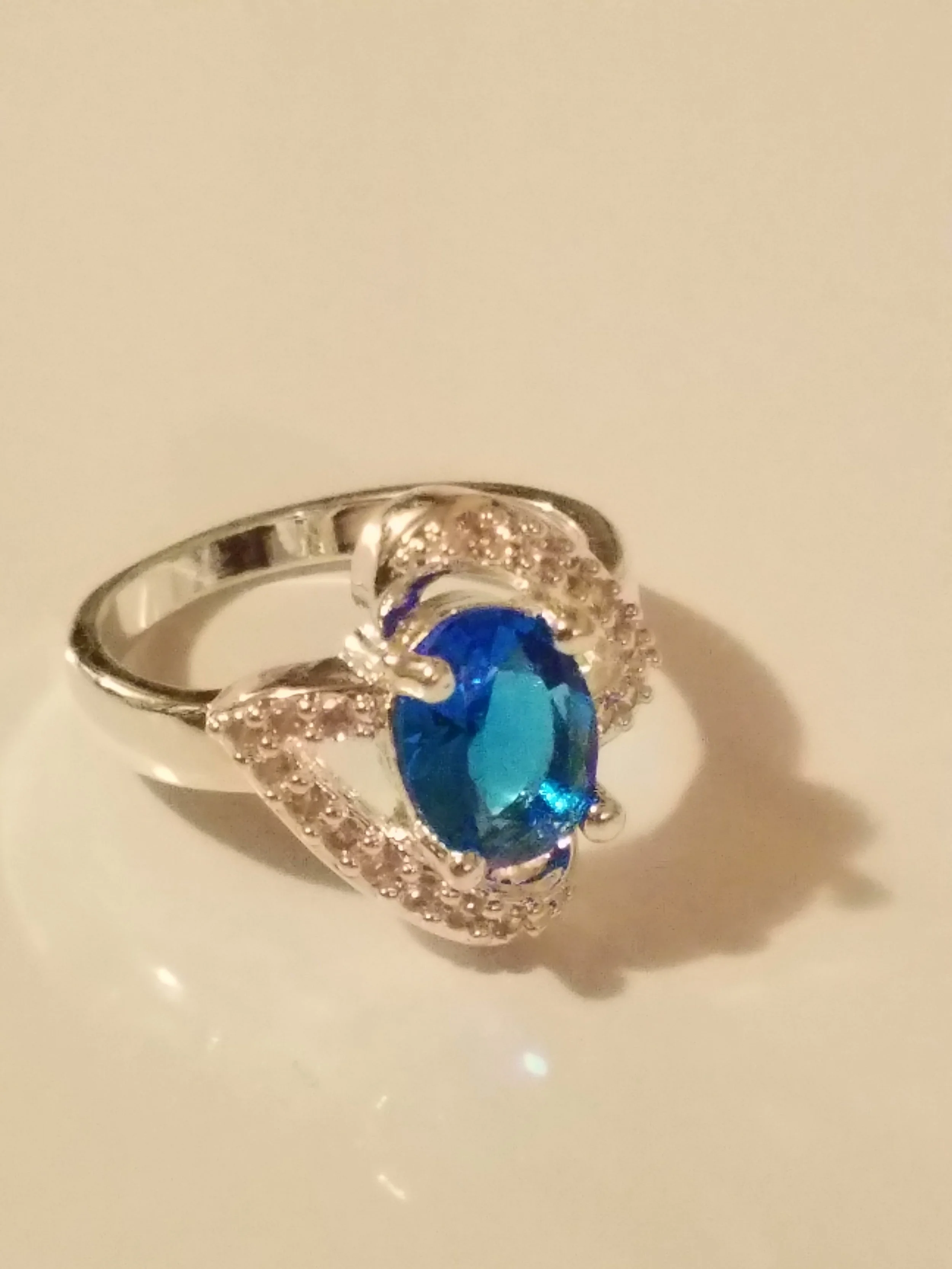 Silver ring with a large oval blue gemstone and small clear stones surrounding it on a white surface.