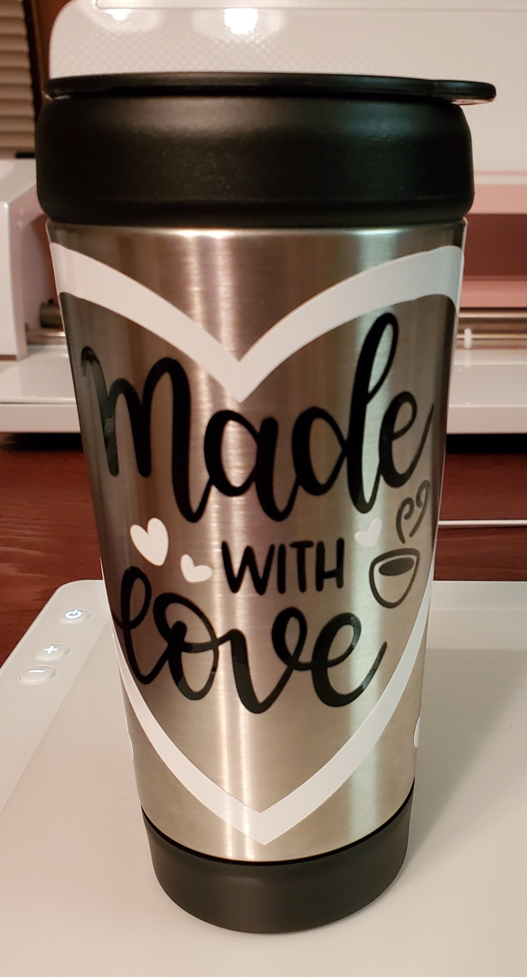 A stainless steel travel mug with a black lid, decorated with a large heart outline and the phrase 'Made with love' written inside the heart, along with small heart and coffee cup illustrations.