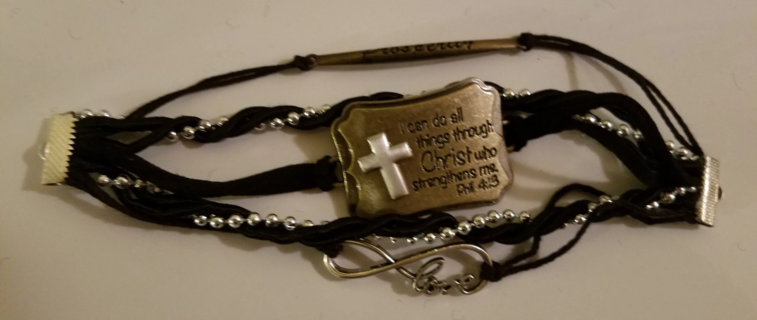 A bracelet made of multiple black cords with silver beads, featuring a rectangular gold-colored metal plate with engraved text and a three-dimensional Christian cross. The text on the plate reads, 'I can do all things through Christ who strengthens m