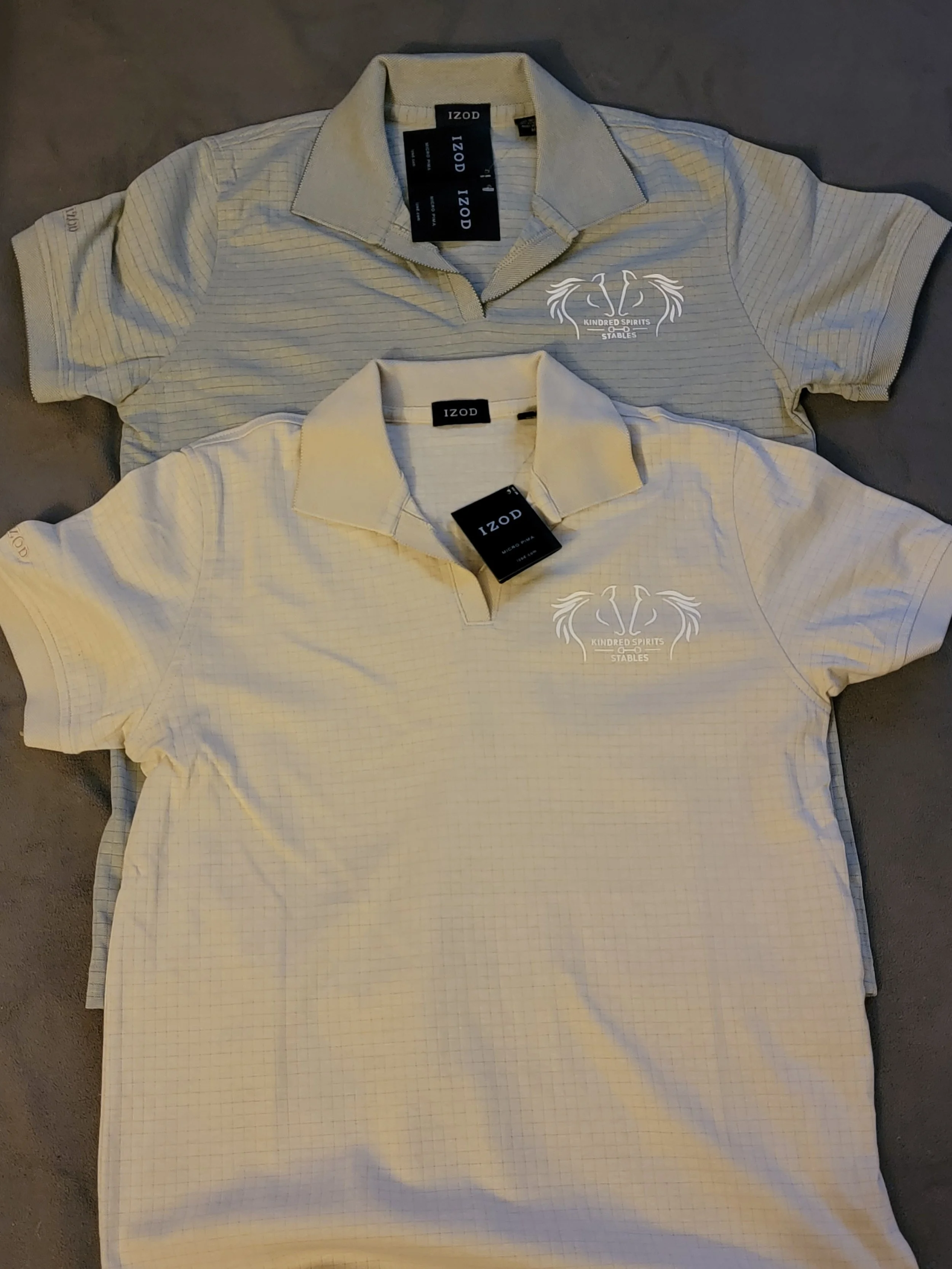 Two IZOD polo shirts, one gray and one cream, laid flat on a surface. Both shirts have a logo with the words "Kindred Spirits Stable" and a horse head design. The shirts have tags attached.