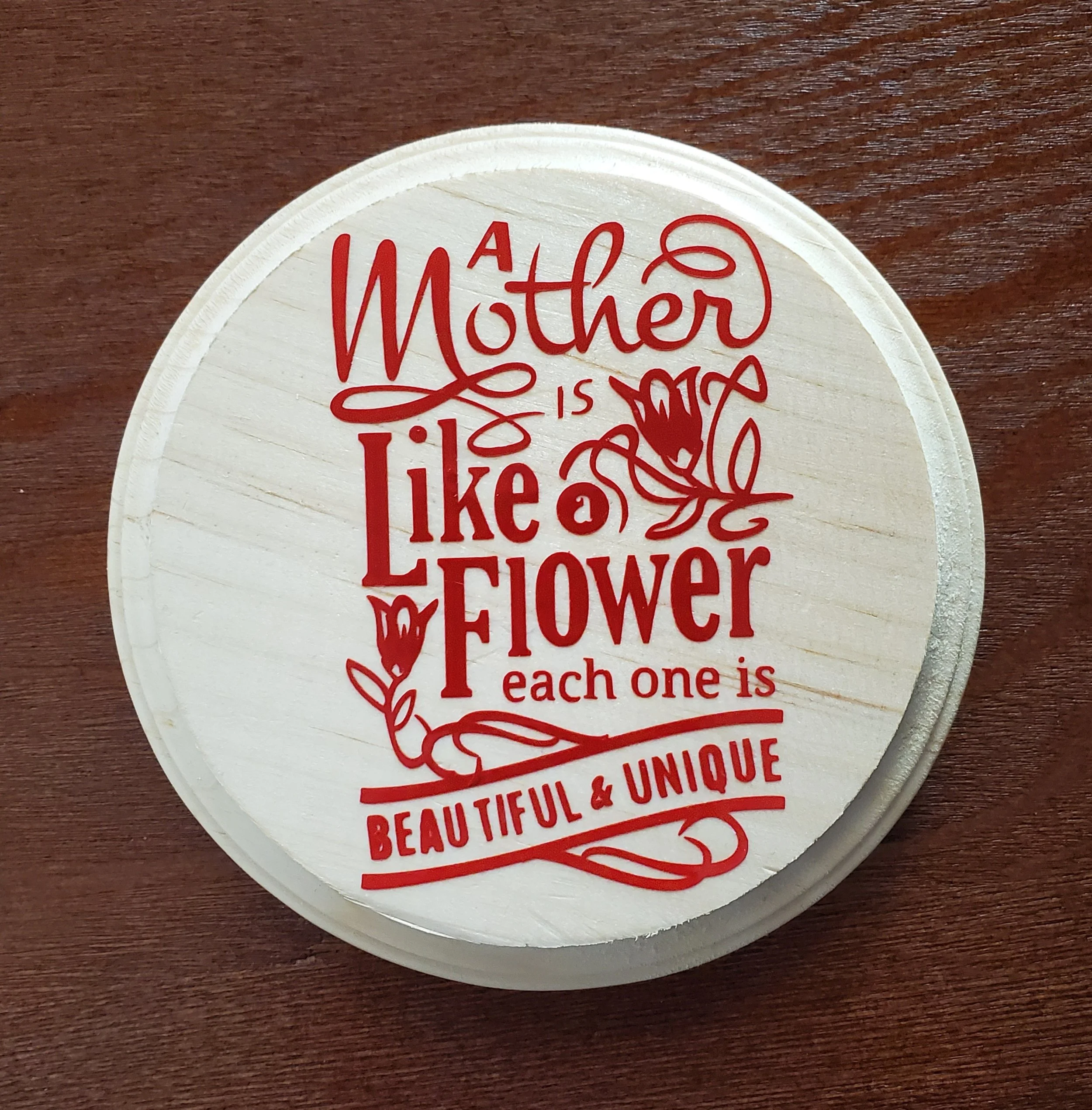 Round wooden plaque with red text that says, "A Mother is like a Flower, each one is beautiful & unique," decorated with floral illustrations.