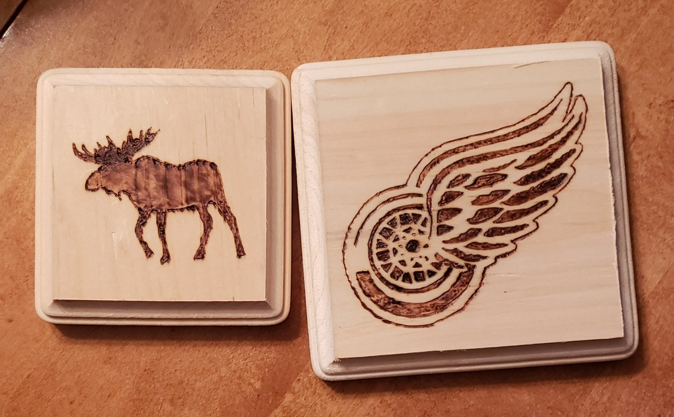 Two wooden blocks with laser-engraved designs: one with a moose and the other with a Detroit Red Wings winged wheel logo.