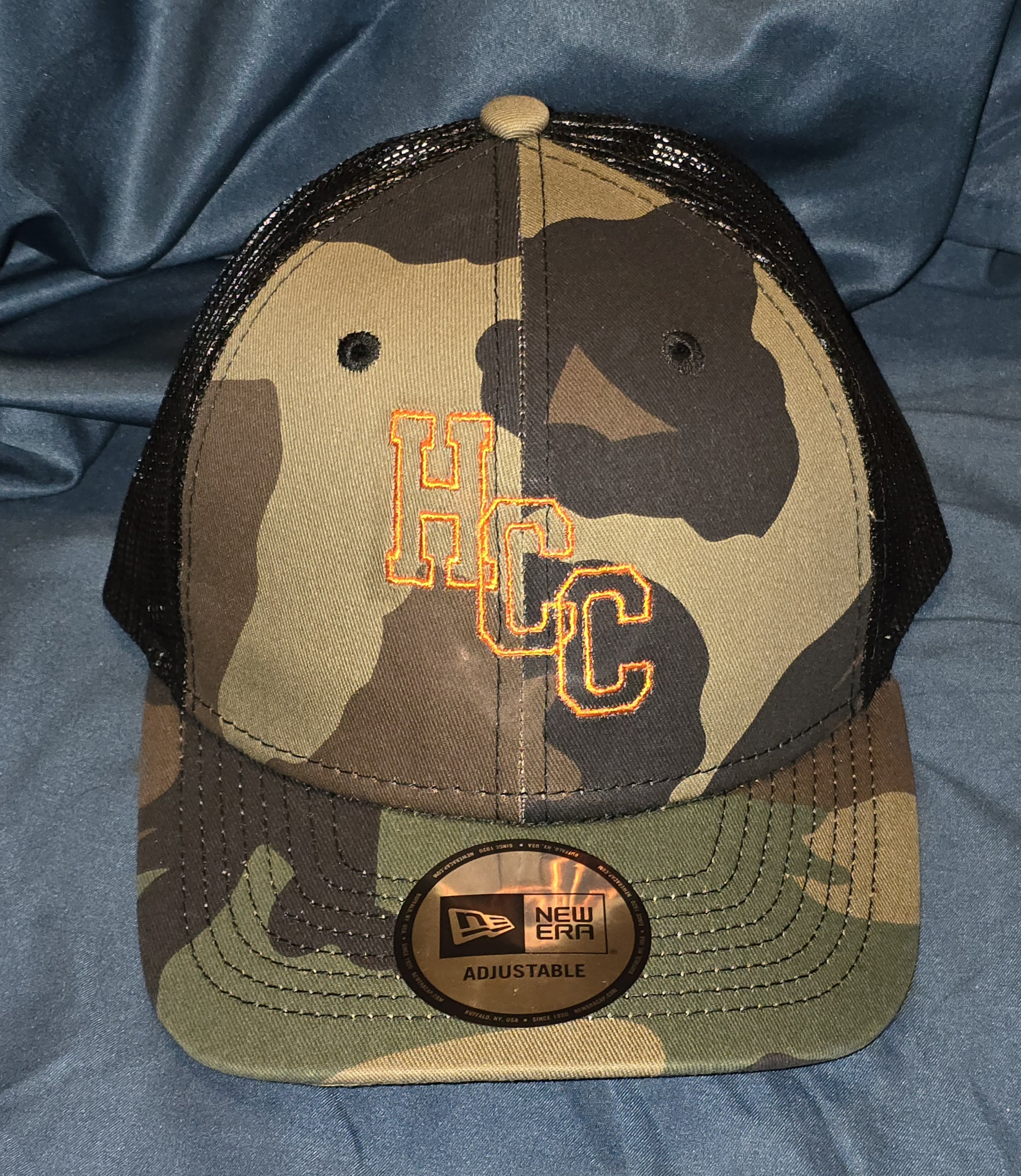 Camouflage baseball cap with embroidered orange letters 'HCC' on front, sitting on a blue fabric background.