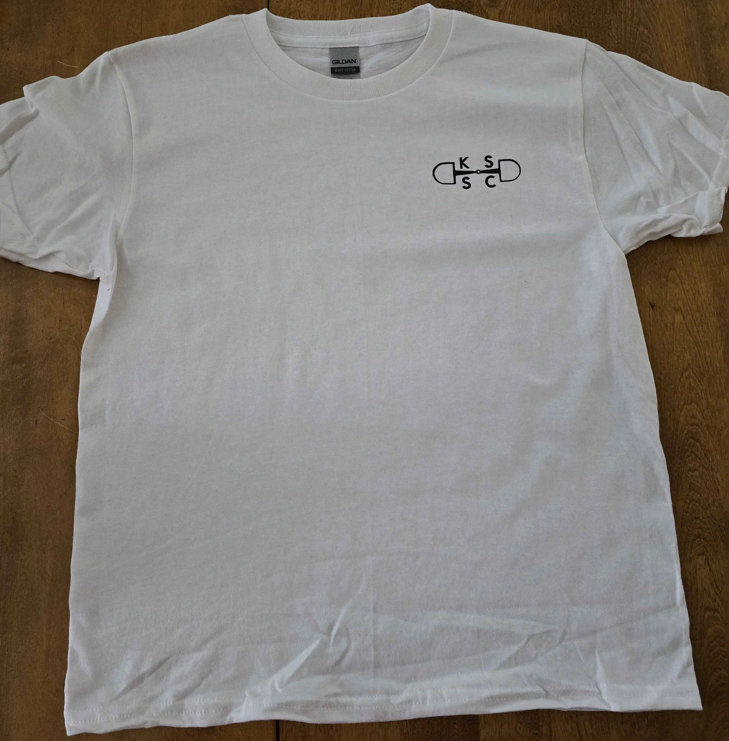 A white Gildan T-shirt with a small black design on the upper left chest area, featuring a capsule pill shape with the letters K, S, C inside it.