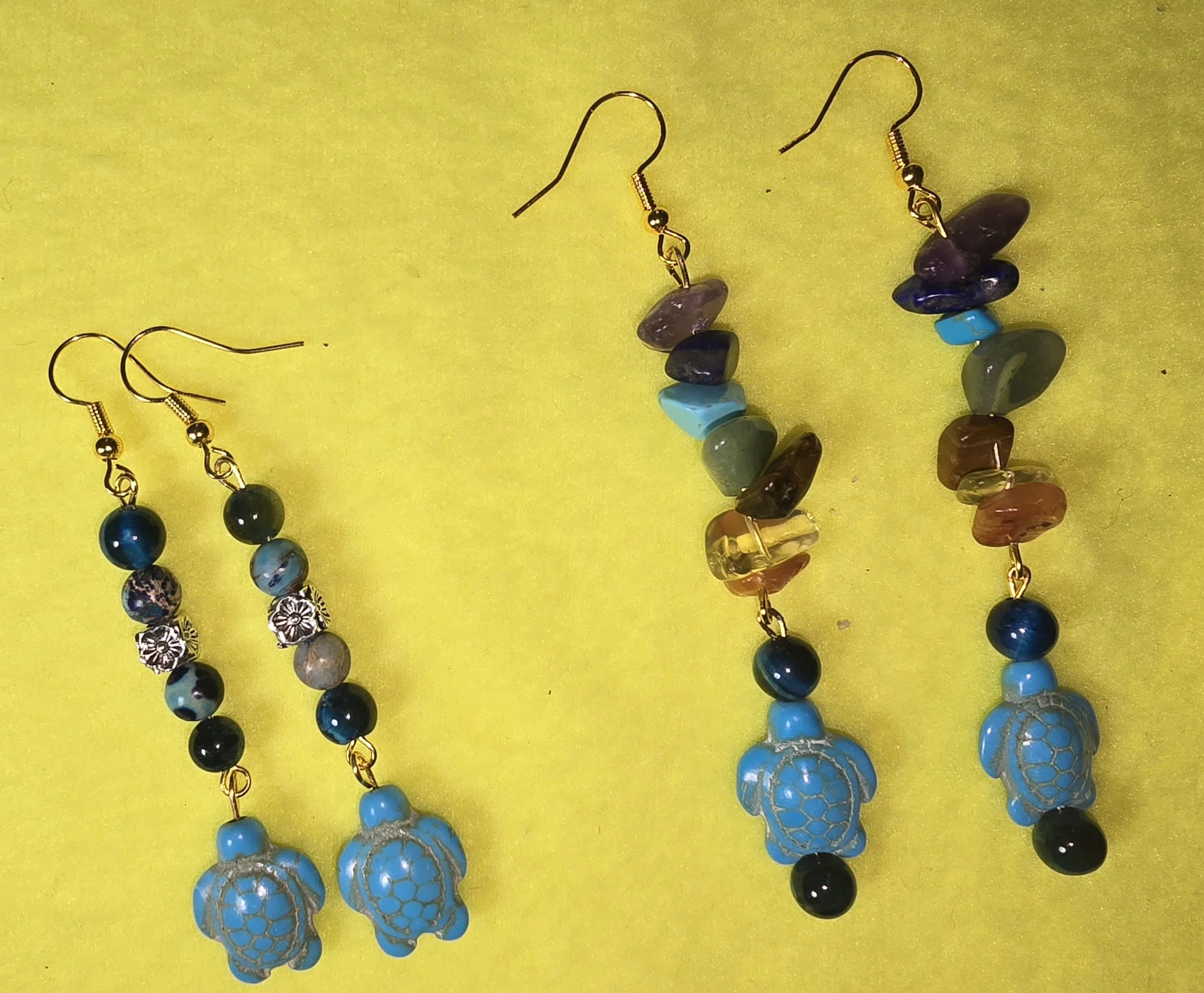 Image of two pairs of handmade earrings with turtle charms, colorful beads, and metal accents, laid on a yellow background.