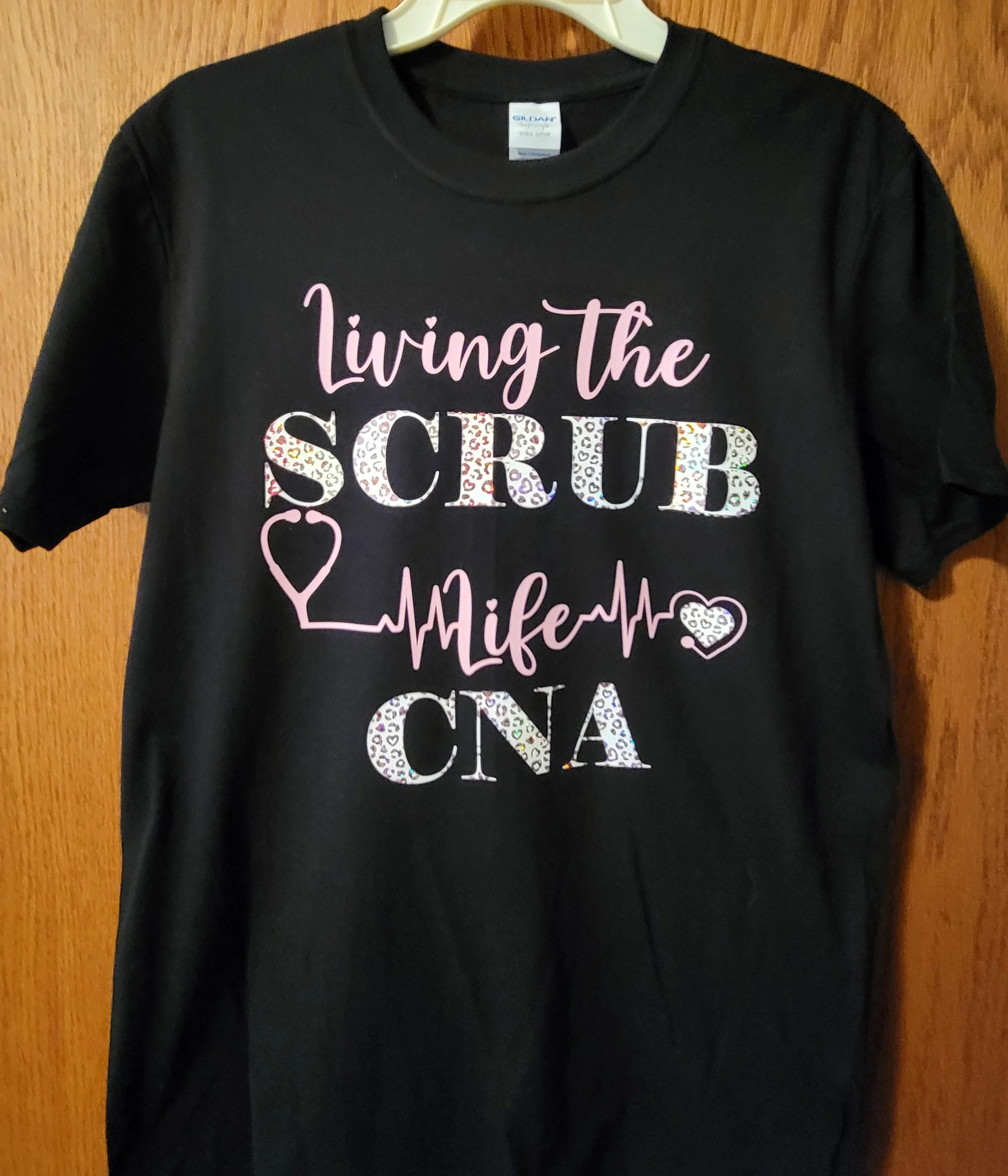 Black T-shirt with pink and white text that says 'Living the Scrub Life CNA' and decorative medical-themed graphics.