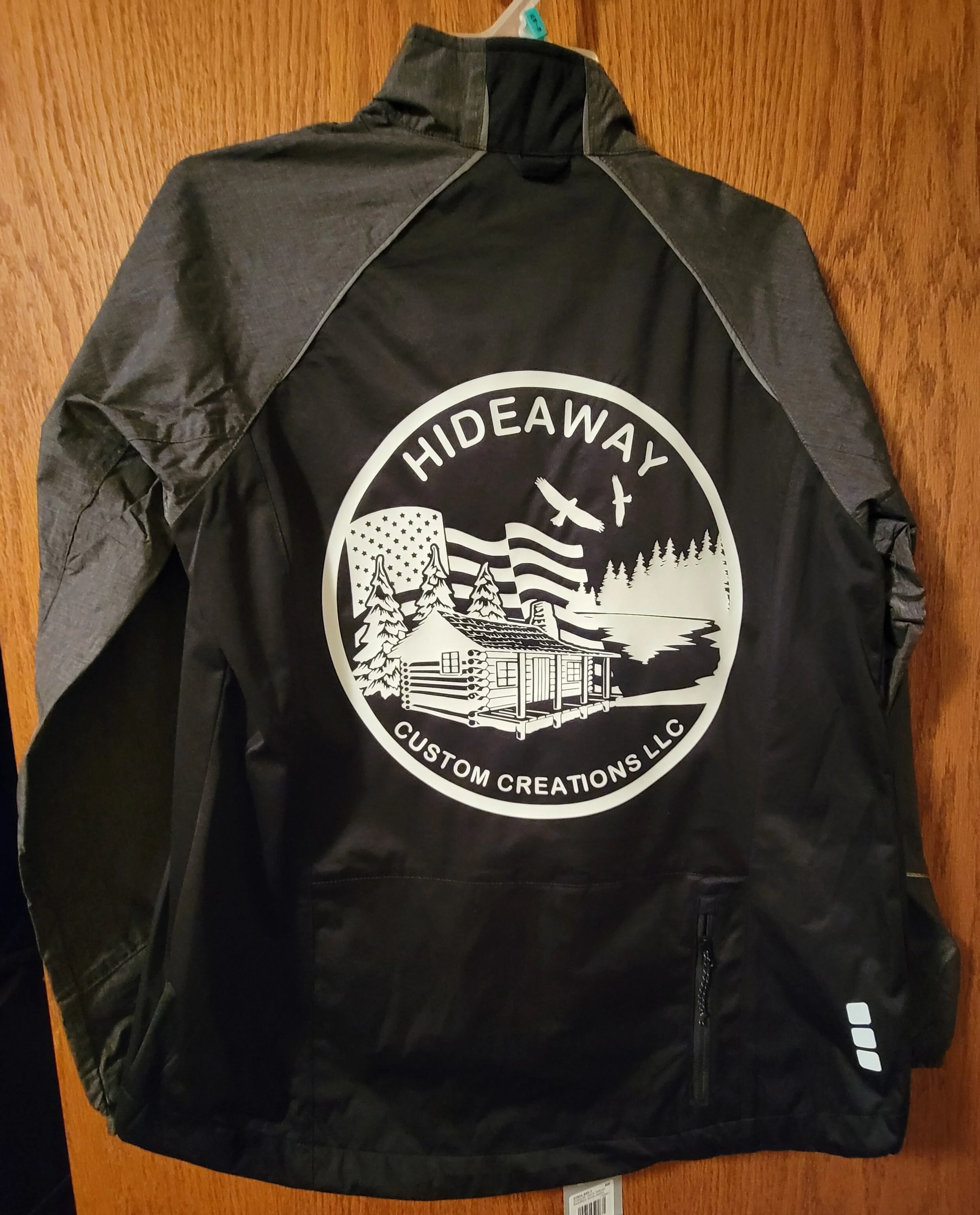Black and gray windbreaker jacket with a large circular logo on the back. The logo depicts a cabin, trees, mountains, and flags with the text 'HIDEAWAY' at the top and 'CUSTOM CREATIONS LLC' at the bottom.