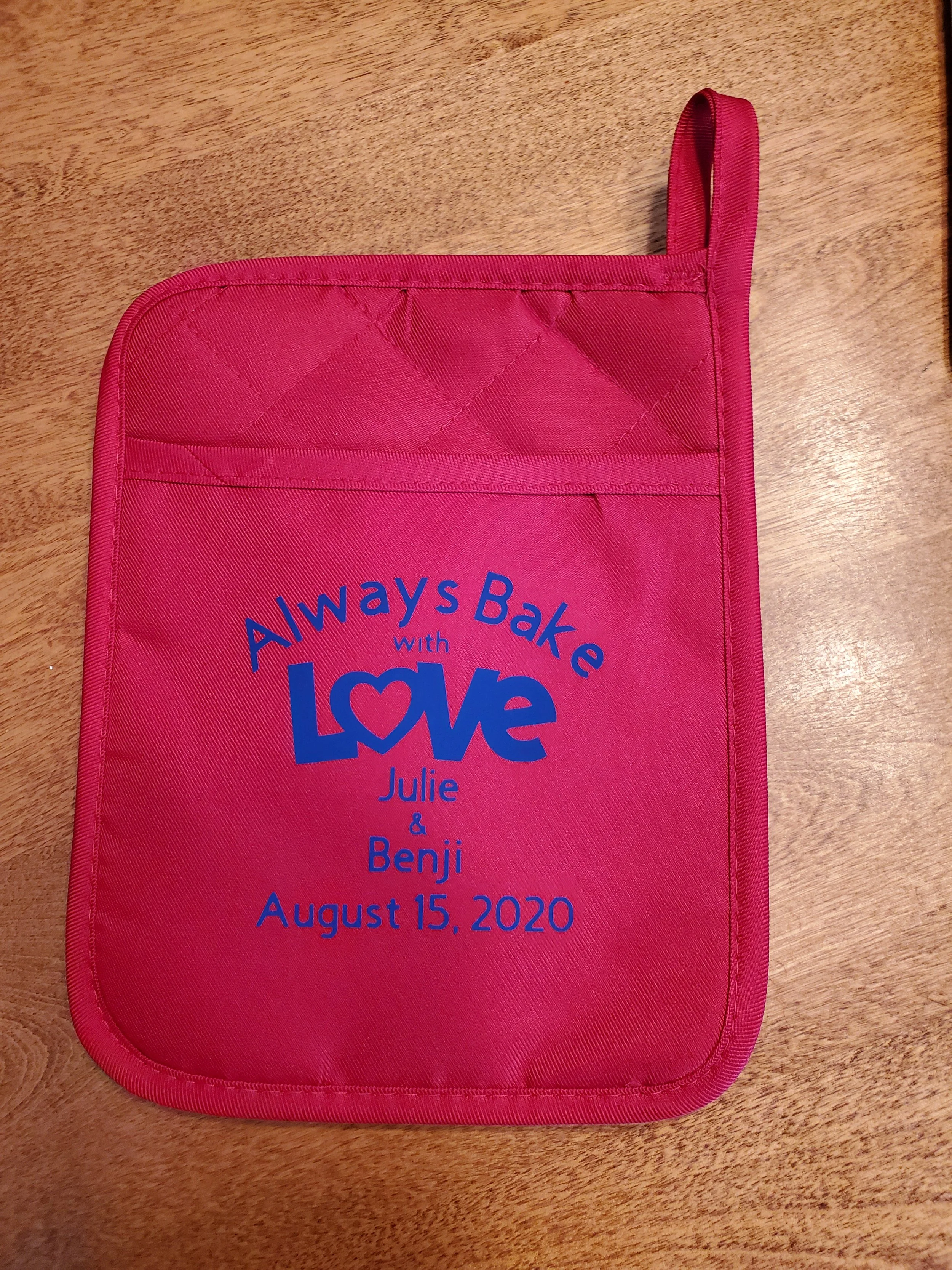 Pink oven mitt with blue text that says, "Always Bake with Love, Julie & Benji, August 15, 2020," on a wooden surface.