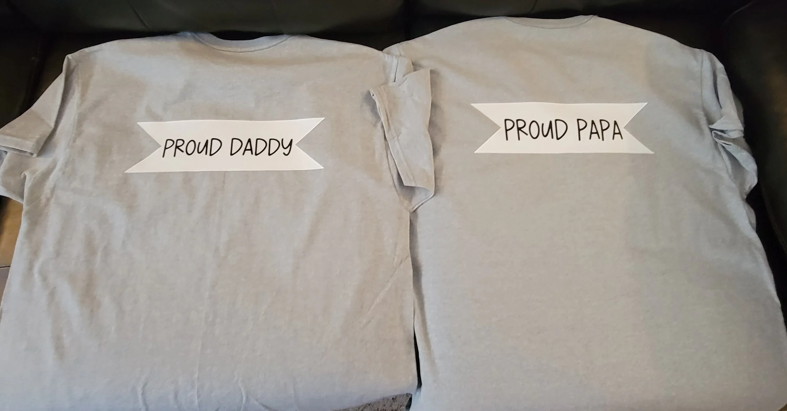 Two gray T-shirts laid flat on a surface, one with a white banner label reading 'PROUD DADDY' and the other with a similar label reading 'PROUD PAPA'.