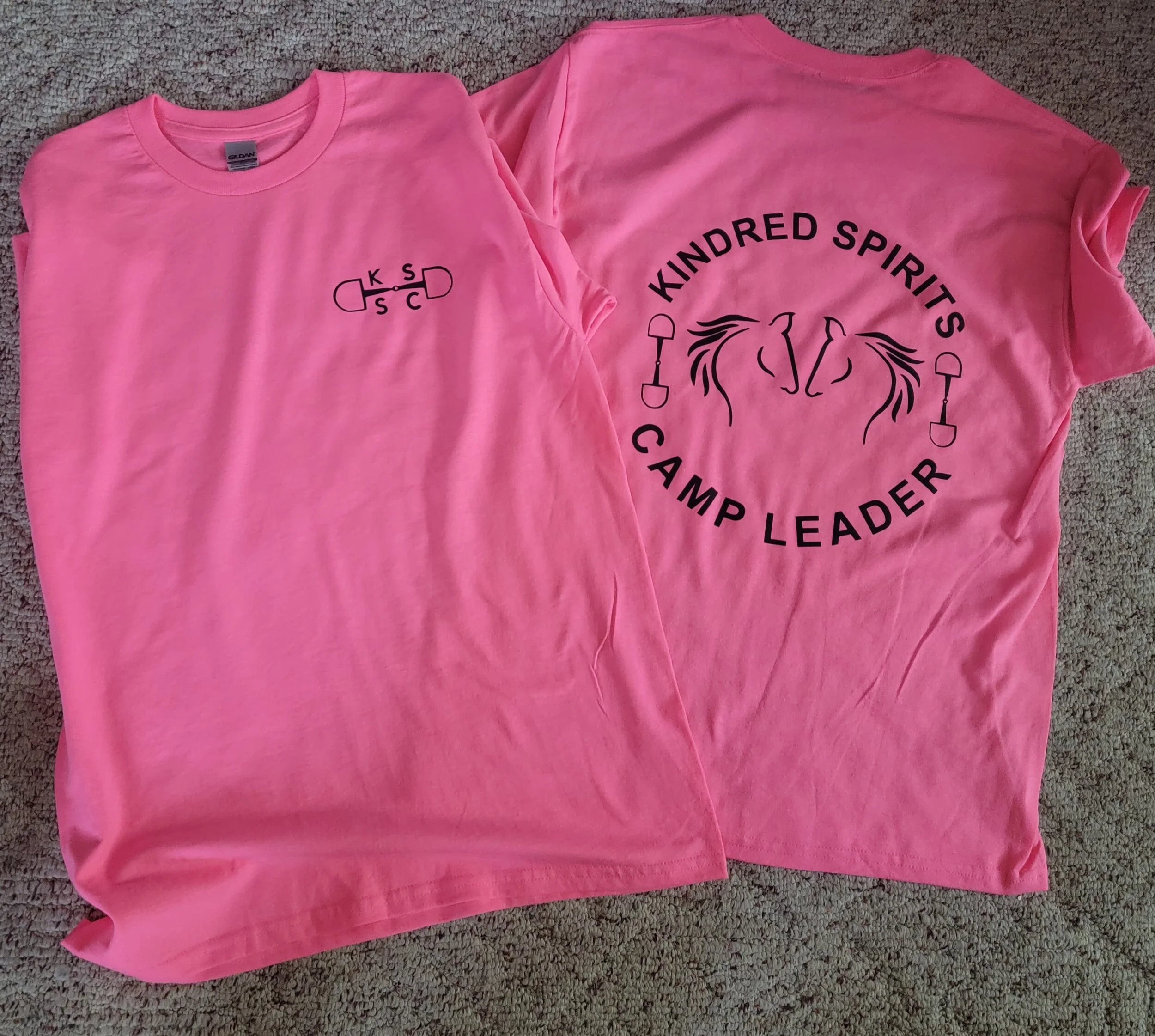 Two pink camp t-shirts laid flat on carpet, one showing the front and the other showing the back. The front has a small logo with an arrow and the letters K, S, D, S, and C, and the back has a larger design with the words 'Kindred Spirits Camp Leader