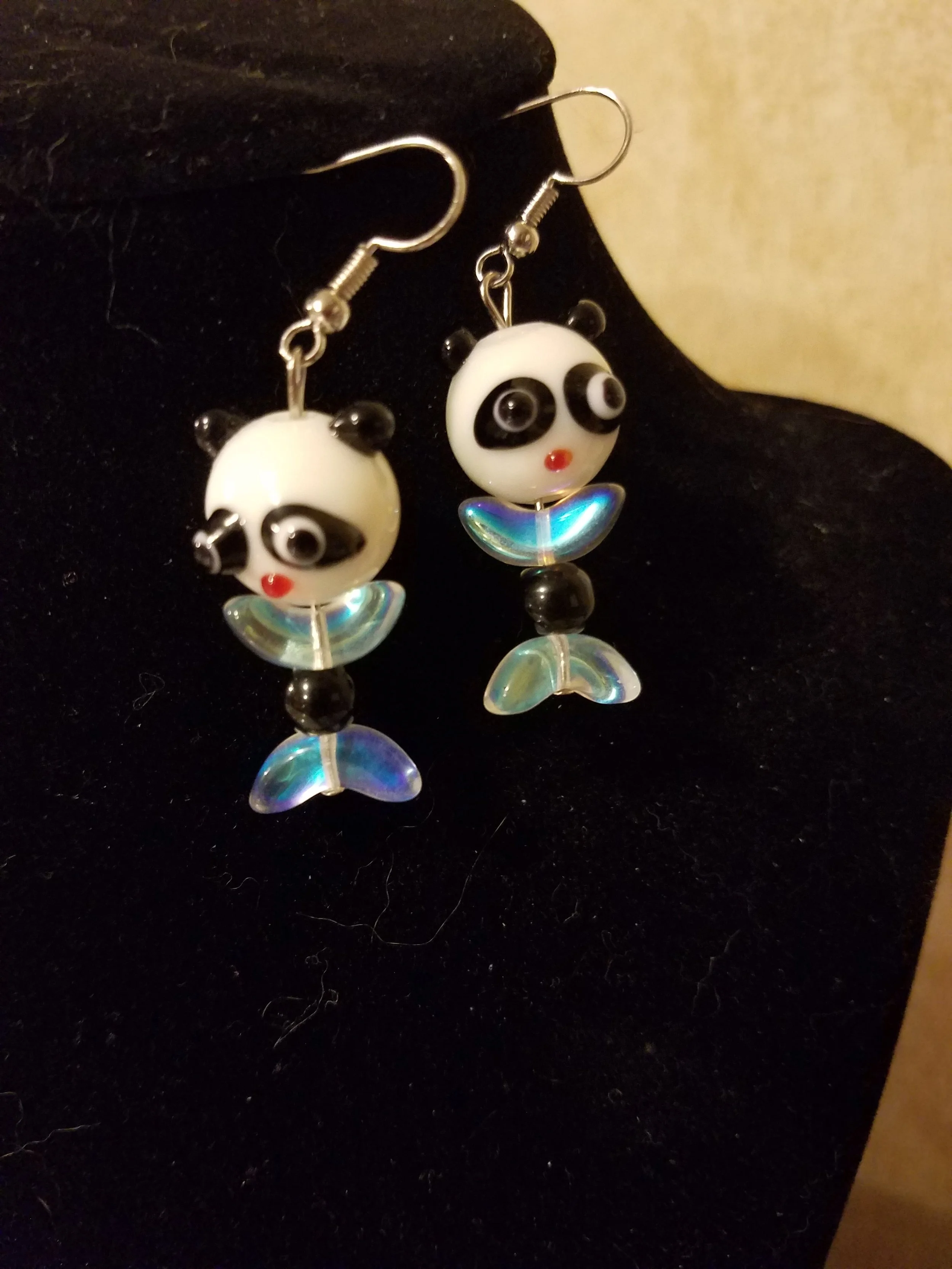 A pair of panda-shaped earrings with black and white panda faces and colorful iridescent tail details, hanging on a black display.