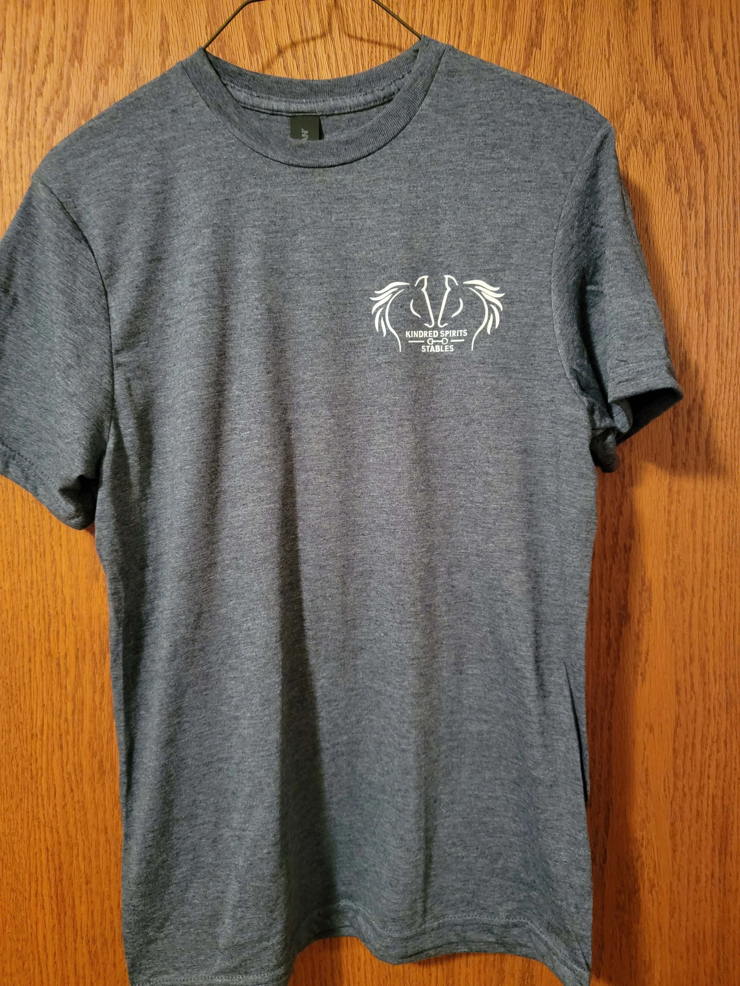 Gray T-shirt hanging on a wooden door with a small logo that reads 'Kindred Spirits & Stables'.