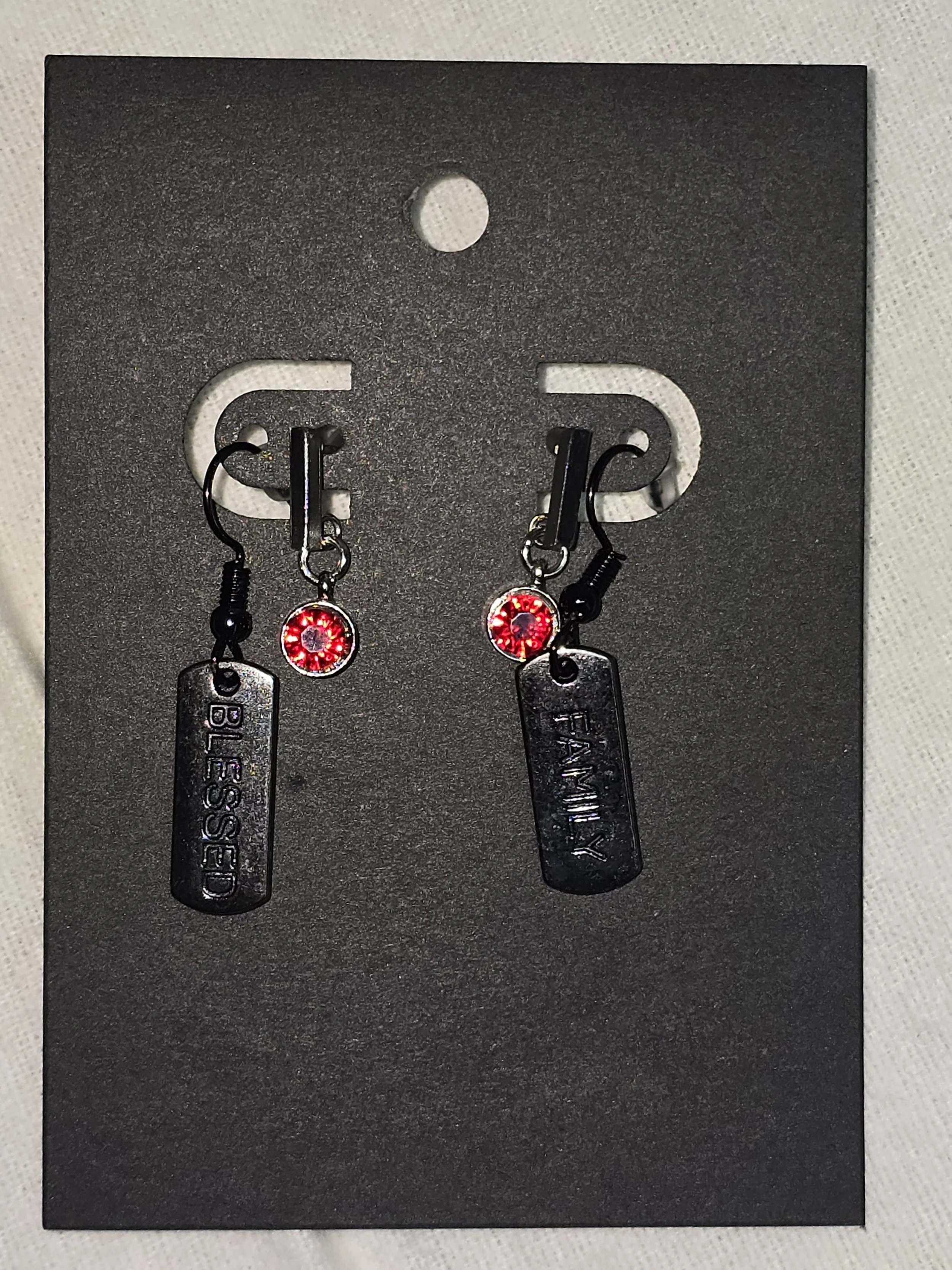Pair of earrings displayed on a black card, one with a tag labeled 'BLESSED' and the other with a tag labeled 'FAMILY'.