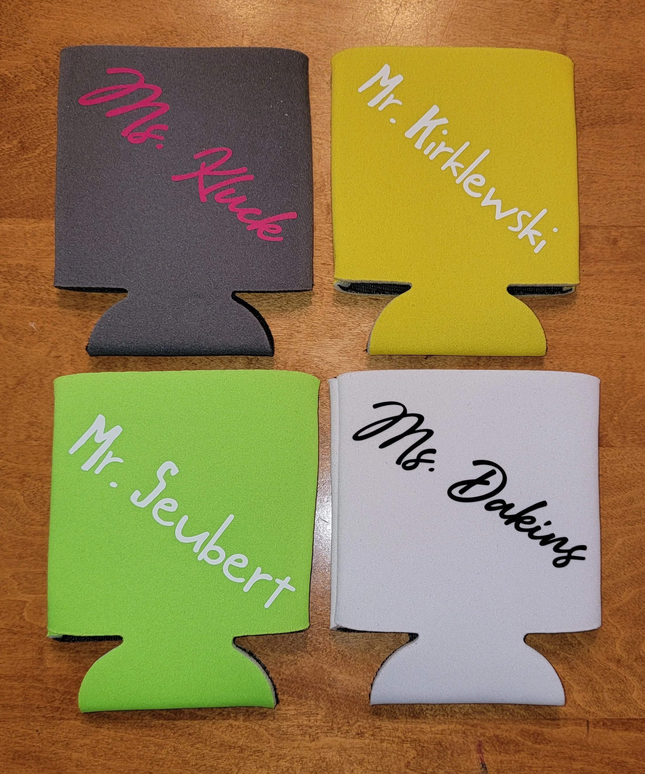 Four colorful foam can cooler drink insulators with handwritten names on each, arranged on a wooden surface. The insulators are purple with pink writing reading 'Ms. Rhiabe,' yellow with white writing 'Mr. Kirlewski,' green with white writing 'Mr. Ce