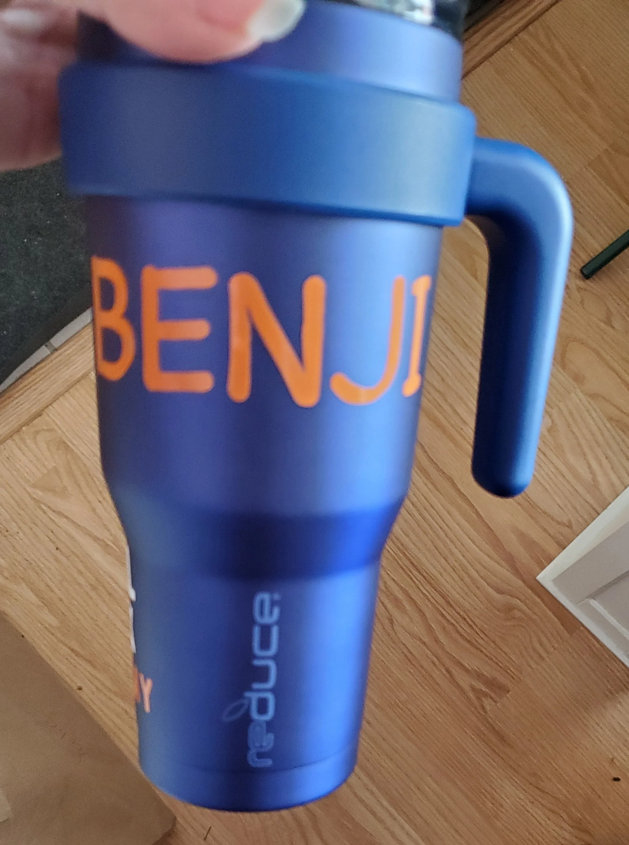 Blue travel mug with orange and white text, reading "BENJ" on the front. The mug is held up by a hand, with a wooden floor in the background.