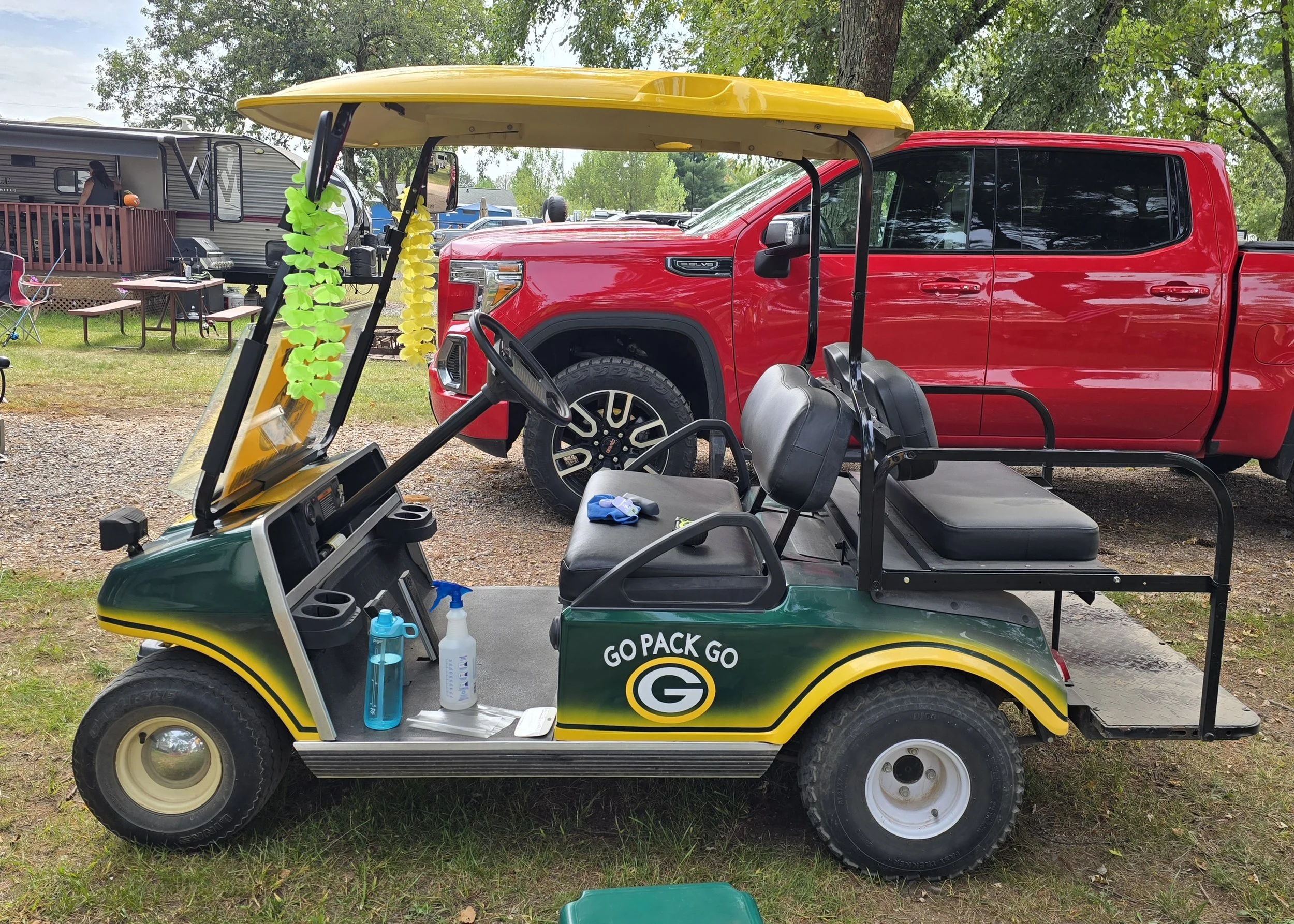 A green and yellow golf cart with bottles of hand sanitizer and spray on the dashboard, parked on grass next to a red pickup truck at a campground with RVs and trees in the background.