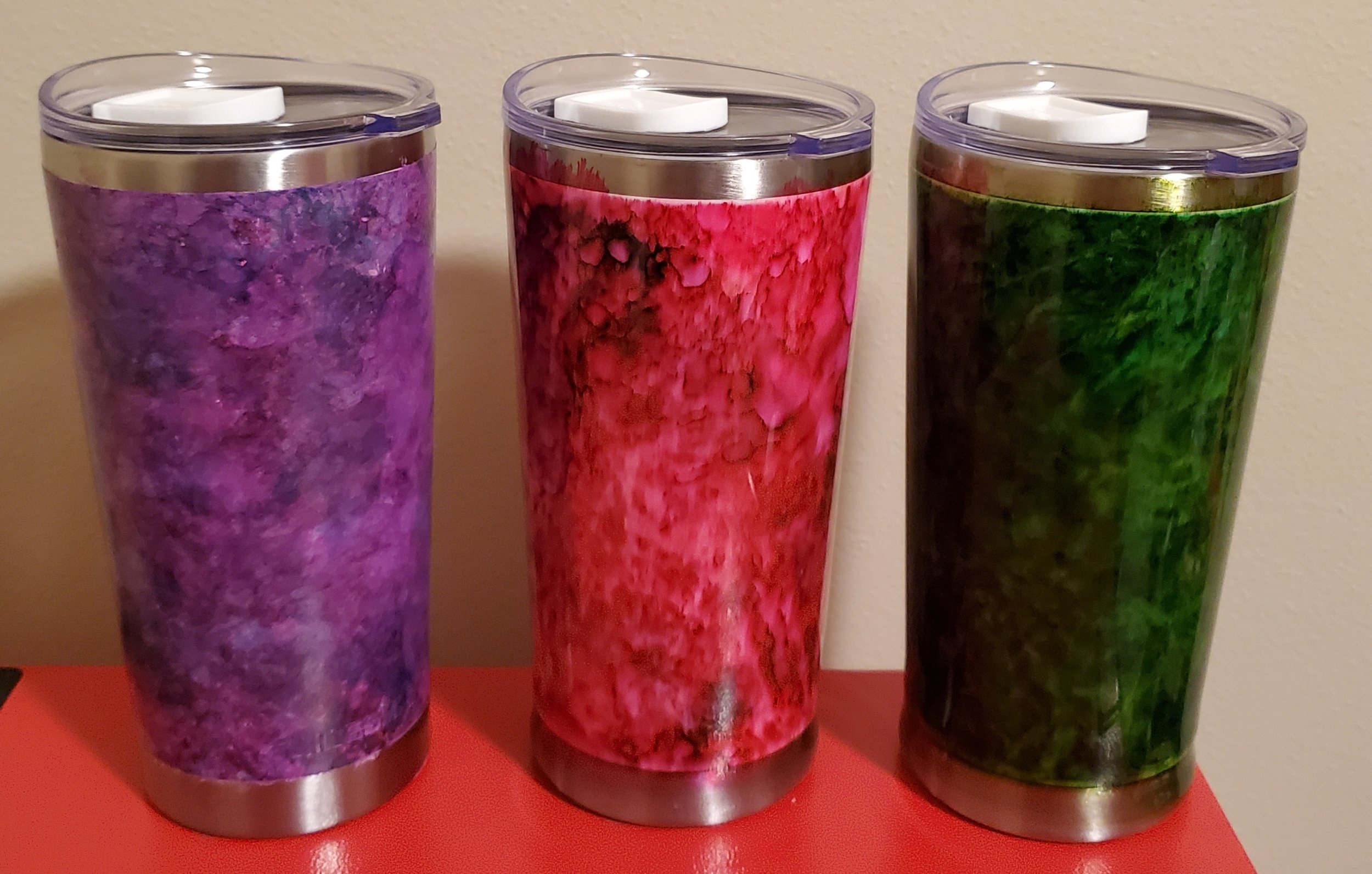 Three stainless steel tumblers with colorful marble-like designs, placed on a red surface against a beige wall.